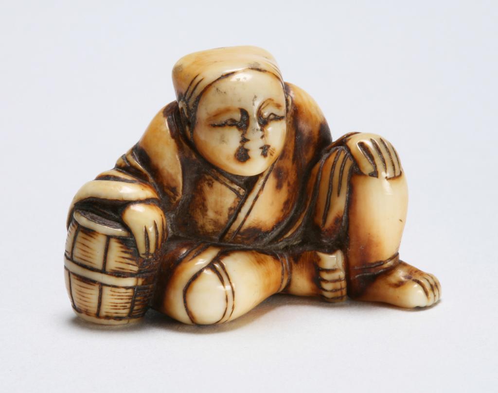An image of Netsuke: katabori: Monkey-trainer. Unknown maker, Japan.  Ivory, carved and stained. Figure of a man (sarumawashi) seated, leaning his right arm on a barrel and resting his left hand on his left knee, his small monkey climbing up his back. Himotoshi on the underside. Ivory, carved and stained, height, whole, 2.9 cm, width, whole, 3.5 cm, circa 1800-1868. Edo Period (1615-1868).