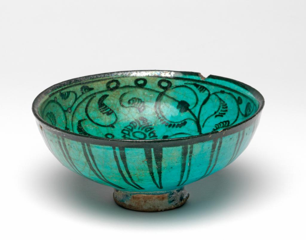 An image of Islamic pottery. Bowl. Unknown potter, Iran. Shape: hemispherical bowl with slightly everted rim, sitting on a slightly flared foot ring. Interior: the rim is painted black. On the body scrolling vines terminate in feathery half palemettes or flower heads; areas close to the rim are adorned with small circles. Turquoise glaze pools in the base. Exterior: on the rim, black paint continues from the interior. On the body pairs of parallel lines radiate from the lower body toward the rim. Dribbles of turquoise glaze cover the foot ring unevenly, pooling in places, over an opaque off-white self-formed? glaze; the latter also coats the underside of the foot, where it is itself partially coated by thin transparent turquoise glaze. Remnants of a thin compact reddish accretion layer coat parts of the exterior glazes, especially the foot exterior and underside with smaller traces on the body. Fritware, wheel thrown. Painted inside and out in black over an opaque off-white self-formed? glaze and under a transparent turquoise glaze. Height, whole, 6.9 cm, width, whole, 14.7 cm, diameter, rim, 14.7 cm, diameter, base, 4.9 cm, circa 1300 to circa 1399. Il-Khanid.
