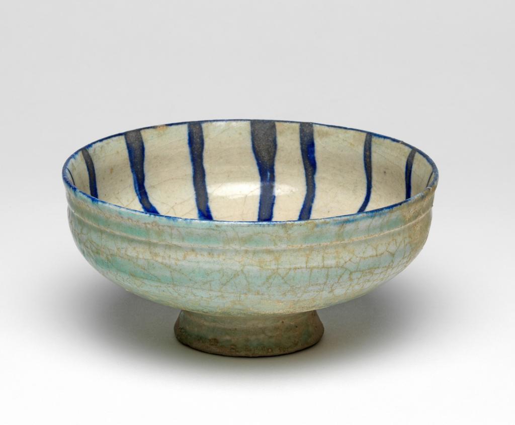 An image of Islamic pottery. Bowl. Unknown potter, Iran. Shape: carinated bowl with a collared neck, sits on a low, flaring foot ring. Interior: painted in dark blue over a white ground beneath a transparent uncoloured glaze. On the rim a blue line is painted. On the body blue lines radiate from a central circle. Exterior: covered with an uneven incomplete glaze with a pale turquoise-green tinge that covers the surface unevenly and terminates above the bottom of the foot ring. Fritware, wheel thrown. Glazed inside and out. Interior painted in dark blue over a white ground under a transparent uncoloured glaze. Exterior glazed with an uneven incomplete glaze with a pale turquoise-green tinge. The underside of the foot is unglazed. Height, whole, 7.5 cm, width, whole, 15.4 cm, diameter, rim, 15.4 cm, diameter, base, 6.3 cm, circa 1200 to circa 1220. Seljuk.