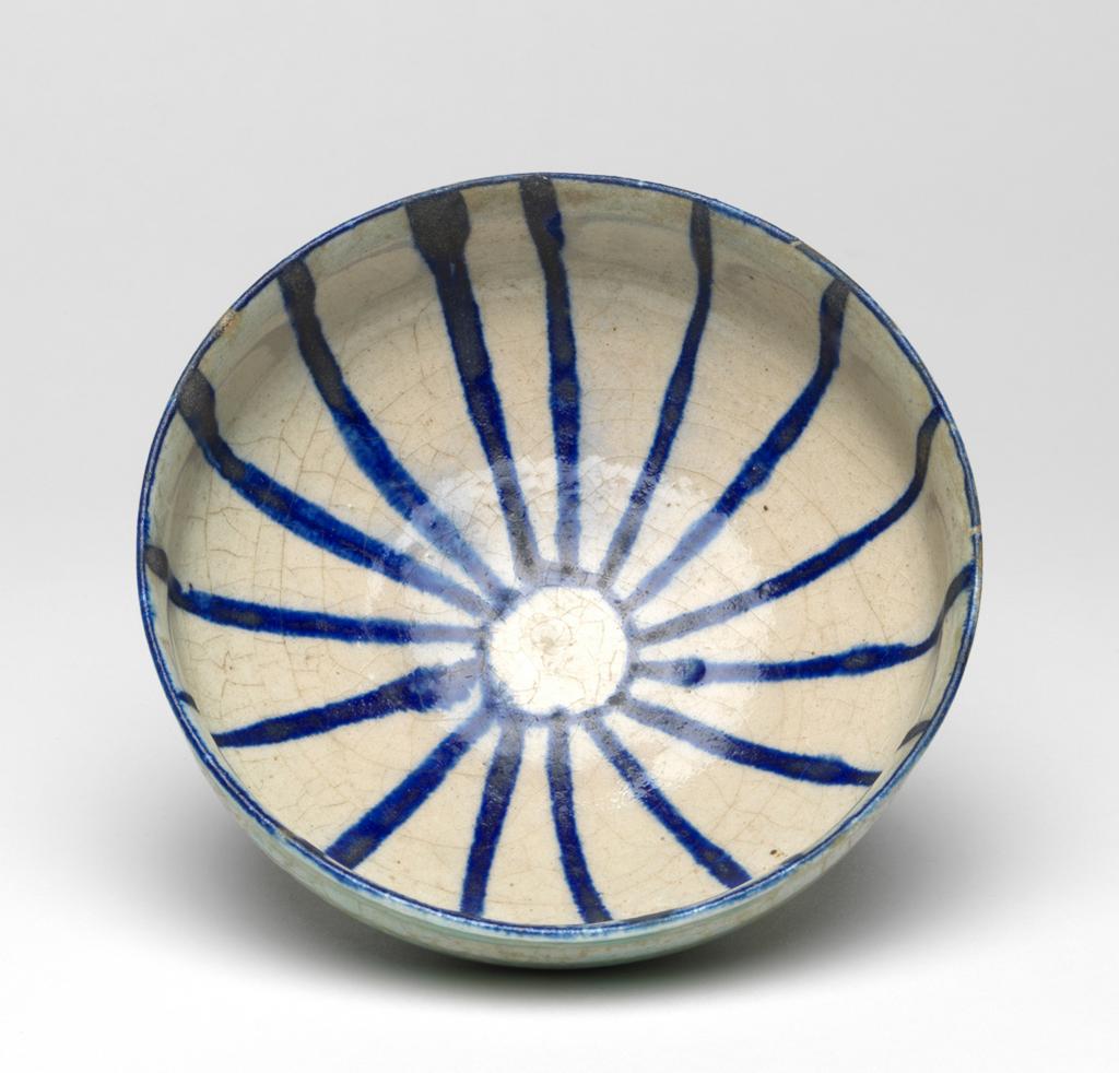 An image of Islamic pottery. Bowl. Unknown potter, Iran. Shape: carinated bowl with a collared neck, sits on a low, flaring foot ring. Interior: painted in dark blue over a white ground beneath a transparent uncoloured glaze. On the rim a blue line is painted. On the body blue lines radiate from a central circle. Exterior: covered with an uneven incomplete glaze with a pale turquoise-green tinge that covers the surface unevenly and terminates above the bottom of the foot ring. Fritware, wheel thrown. Glazed inside and out. Interior painted in dark blue over a white ground under a transparent uncoloured glaze. Exterior glazed with an uneven incomplete glaze with a pale turquoise-green tinge. The underside of the foot is unglazed. Height, whole, 7.5 cm, width, whole, 15.4 cm, diameter, rim, 15.4 cm, diameter, base, 6.3 cm, circa 1200 to circa 1220. Seljuk.
