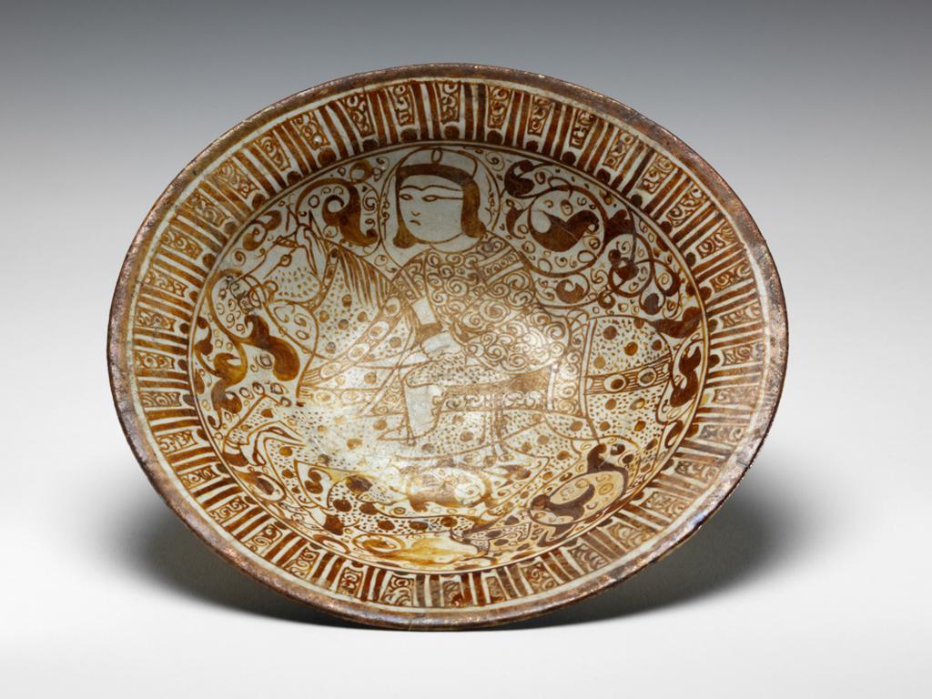 An image of Dish. Miniature style. Islamic pottery. Unknown potter, Iran, probably Kashan. Shape: dish with a flange rim and a low foot ring. Interior: a concentric band covers the rim, below a frieze of pseudo-calligraphy comprising vertical lines and dots interspersed with spirals. The central decoration comprises a dog and mounted horse whose harness is decorated with a peacock’s eye pattern and blanket is decorated with scrolls. The horse rider wears a headdress and a tunic decorated with spirals over trousers with a pattern of triangular dots. Both the body of the horse, and dog, are covered with large dots and small dashes. Scrolling vegetation, including solid palmettes, decorate the background. All decoration is painted in lustre. Exterior: on the body, a frieze of simple repeating arabesques, painted with a single stroke, are separated by pairs of vertical lines and enclosed by concentric lines. Both are painted in lustre. Glaze runs over the body to a point just above or just below the start of the foot ring. Buff fritware, wheel thrown, with an opaque white, slightly crazed glaze and yellow-brown lustre painted decoration. Height, whole, 5.7 cm, diameter, rim, 23 cm, diameter, base, 9.2 cm, circa 1179-1198. Seljuk. Acquisition: bequeathed 1950; Henry Scipio Reitlinger.