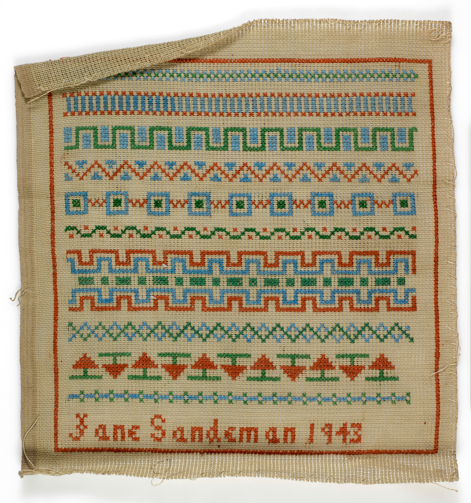 An image of Embroidery Sampler. J.J. Sandeman. Double weave canvas embroidered with polychrome wools in cross stitch. A single line of cross stitch frames 10 horizontal bands of different repeat patterns, inscribed "J.J.Sandeman 1943" at the bottom. 1943.