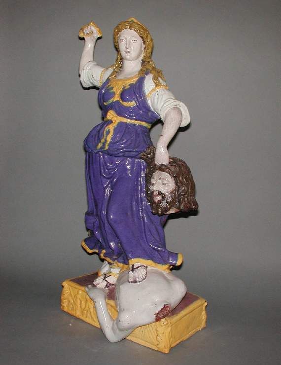 An image of Maiolica Figure Group. Judith and Holofernes. Della Robbia, Giovanni, imitation of. Judith stands on a rectangular pedestal, moulded in relief with an acanthus leaf on each front corner and with a frieze of figures across the front. Her weight is on her right foot and she rests her left on the torso of Holofernes, whose left shoulder and arm project over the edge of the pedestal. Judith holds his decapitated head by its hair in her left hand; her right arm is raised, the hand grasping a sword hilt (the blade missing). She wears a long gown with elbow-length sleeves, and a full-skirted, sleeveless tunic, fastened by a belt and doubled up over her hips. She has sandals, a bead armlet on each arm and a small tiara or frontlet. Pale buff terracotta, tin-glazed on all surfaces except the back of the pedestal and interior, which is hollow. Glaze colours are blue, yellow, brownish-yellow, brown, manganese-purple and red. Height, whole, 62.0 cm, width, base at front, 28.0 cm, depth, base, 21.5 cm, circa 1800-1860. Renaissance style.