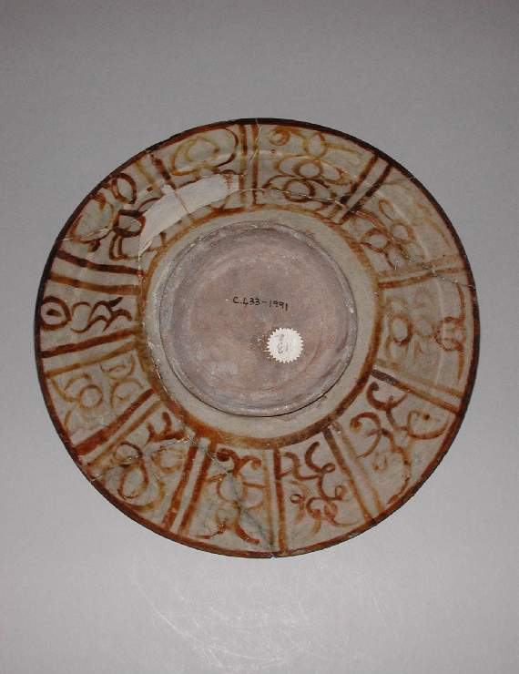An image of Dish. Miniature style. Islamic pottery. Unknown potter, Iran, probably Kashan. Shape: dish with a flange rim and a low foot ring. Interior: a concentric band covers the rim, below a frieze of pseudo-calligraphy comprising vertical lines and dots interspersed with spirals. The central decoration comprises a dog and mounted horse whose harness is decorated with a peacock’s eye pattern and blanket is decorated with scrolls. The horse rider wears a headdress and a tunic decorated with spirals over trousers with a pattern of triangular dots. Both the body of the horse, and dog, are covered with large dots and small dashes. Scrolling vegetation, including solid palmettes, decorate the background. All decoration is painted in lustre. Exterior: on the body, a frieze of simple repeating arabesques, painted with a single stroke, are separated by pairs of vertical lines and enclosed by concentric lines. Both are painted in lustre. Glaze runs over the body to a point just above or just below the start of the foot ring. Buff fritware, wheel thrown, with an opaque white, slightly crazed glaze and yellow-brown lustre painted decoration. Height, whole, 5.7 cm, diameter, rim, 23 cm, diameter, base, 9.2 cm, circa 1179-1198. Seljuk. Acquisition: bequeathed 1950; Henry Scipio Reitlinger.