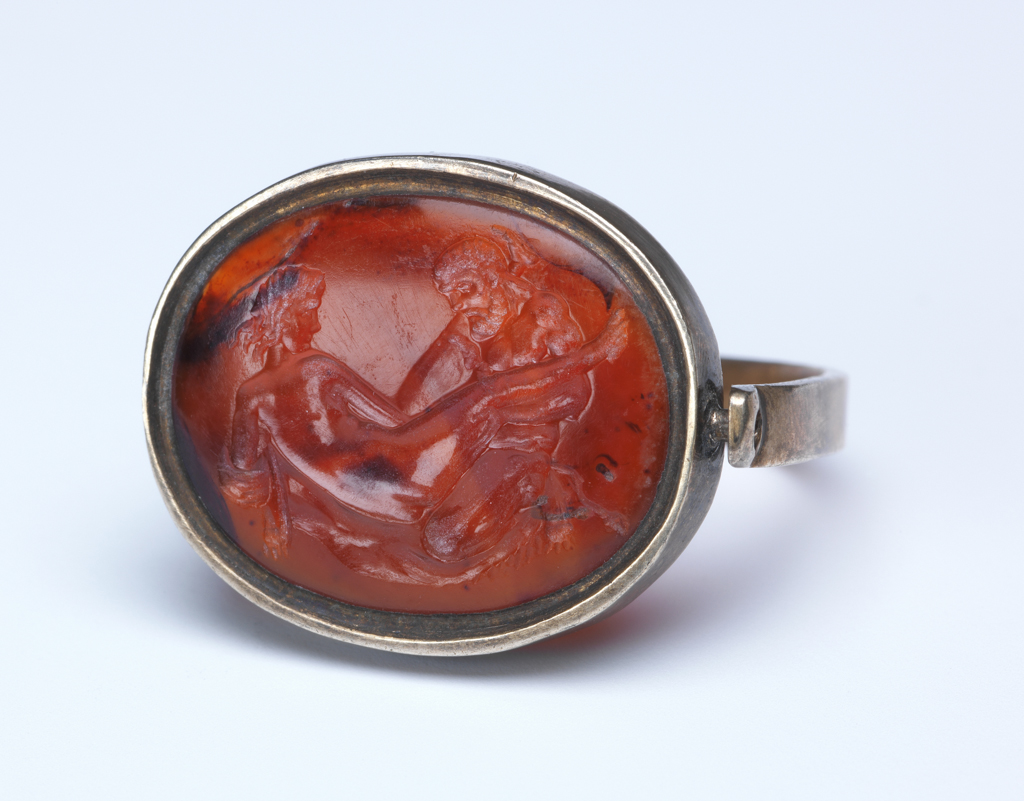 An image of Engraved gems/sealstone/intaglio. A love-scene, with Hercules and Omphale on a bed covered with the lion-skin. Mount: silver swivel-ring, type 3. Intaglio cutting, sard, height 19 mm, width 15.5 mm, circa 1500- circa 1700.