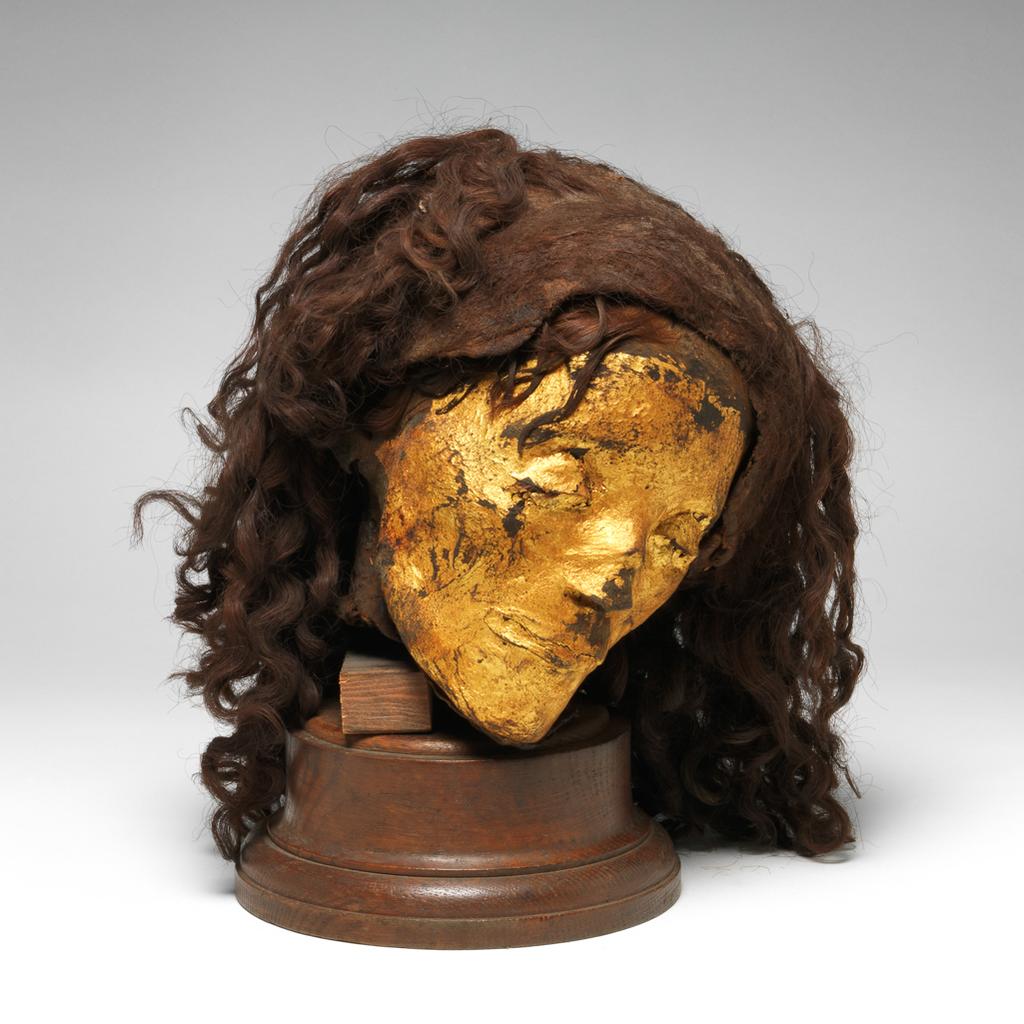 An image of Organic remains. Human mummy. Female head, mummified, with gilded skin, with wig (hair length 0.320 m). Production Place/Find Spot: Egypt. Depth 0.21 m, height 0.22 m, width 0.19 m.