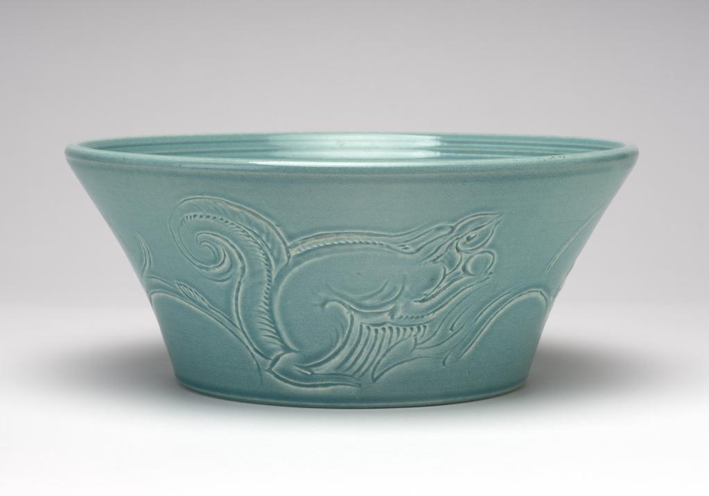 An image of Bowl. Cooper, Susie (British, 1902-1995). Wood & Sons Ltd (Maker). Bowl with wide flat base and flared, slightly concave sides. On the outside, three carved designs depicting a squirrel, one a reversed version of another, the third different. On the inside, three incised lines around the bowl near the rim, visible throwing lines on the base. Slightly mottled jade green matt glaze. Underside flat and glazed within foot rim. Earthenware, moulded, carved and glazed jade green. Diameter 24 cm, height 10 cm. Circa 1932-1933.
