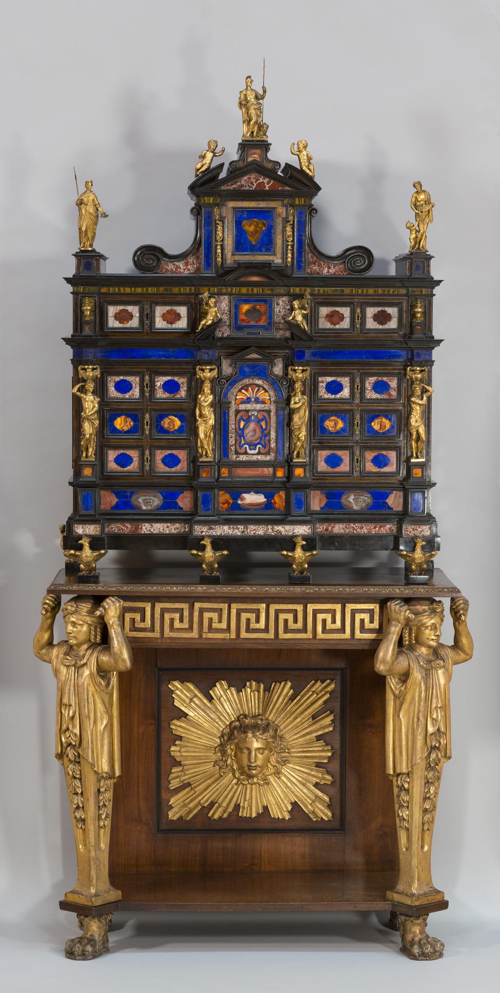 An image of Furniture/cabinet on stand. Rosewood cabinet, mounted with pietre dure, and inlaid with ebony. On a Regency mahogany and parcel-gilt stand. One of a pair. Unknown, maker, Italy, Rome, Tatham, Charles Heathcote, designer, possibly (British, 1772-1842). The cabinet is of 'architectural' form. The upper part has pediments and drawers, and has a central cupboard which encloses a fitted interior with an arcaded alcove. The pediments are surmounted by classical figures and putti, who stand flanked by female figures, with summer and autumn attributes. The cabinet is inset with panels of lapis lazuli, agate, quartz, and various Roman marbles. The pilasters are formed of four gilt bronze standing figures, with gilt bronze capitals with rams heads, on plinths supported by four gilt bronze crowned eagles. The stand is on lion paw feet, and has a moulded top and gadrooned edge above a central frieze, with Greek key decoration, above two parcel gilt caryatid supports with floral drop swags. On the back panel is a central plaque, with a female mask, centrered by gilt wood sun rays. Overall height 222 cm, width 92 cm, depth 43.5 cm, cabinet circa 1625, stand first quarter 19th century. Baroque. Production Note: This cabinet is a pair with M.13 & A-2016. They were made for a member of the Roman Borghese family about 1625, and were later mounted on English neo-classical stands and displayed as Grand Tour trophies in the Long Gallery at Castle Howard.