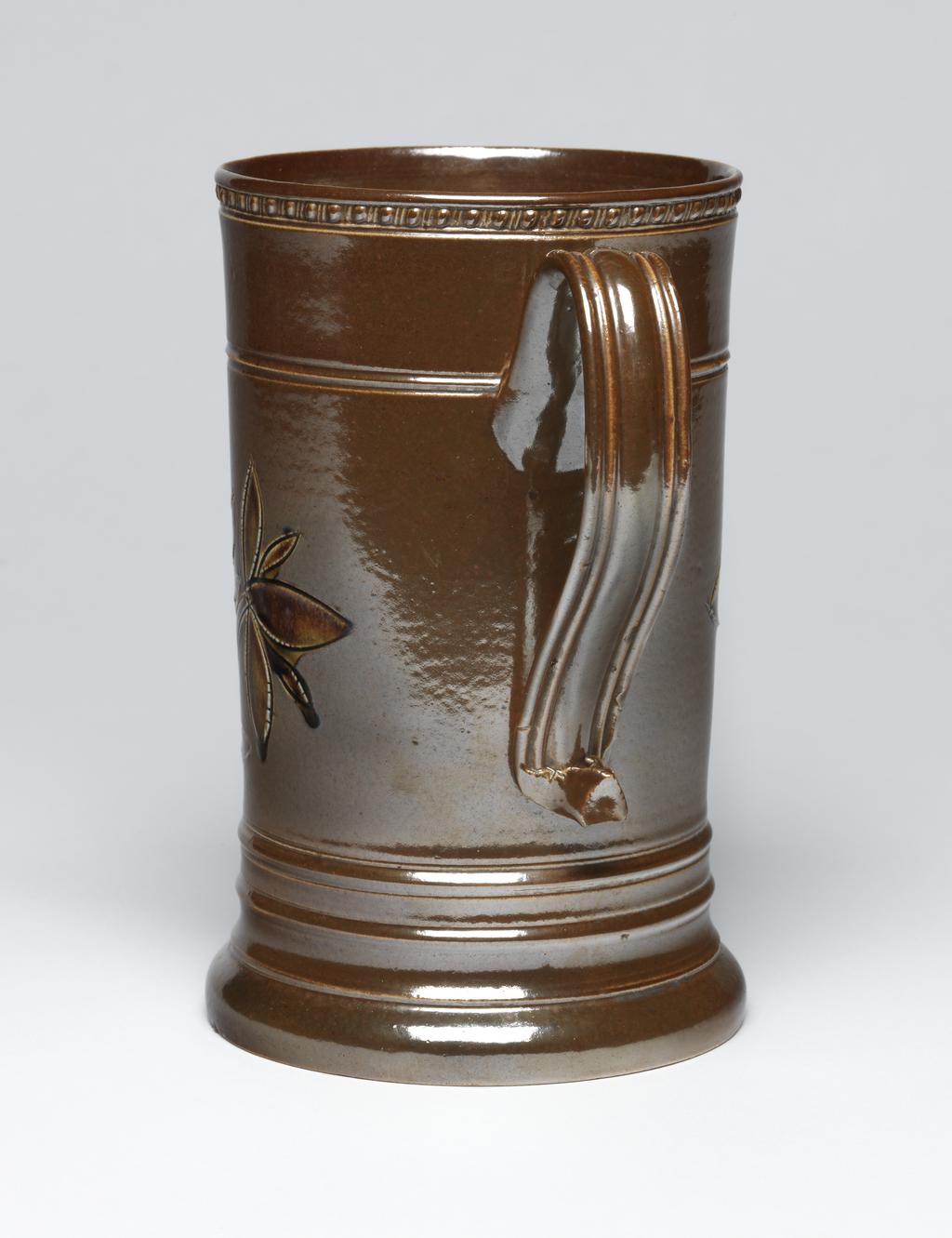An image of Tankard/Mug. Unidentified Nottingham pottery, England. Cylindrical with slightly projecting base, two projecting bands below the handle and one level with its junction with the body at the top, and a row of oval beading round the rim. The ear-shaped handle is reeded and has an upward kick at the bottom. The front is incised with a flowering bush painted with dark brown slip. Four stilt marks on base. Brown salt-glazed stoneware, thrown, turned, rouletted, incised and painted with dark-brown slip. Height, whole, 18.0 cm, diameter, base, 11.3 cm, width, whole incl handle, 14.5 cm, circa 1750-1770. Production Note: Incised and slip-painted decoration is the rarest kind of decoration on Nottingham salt-glazed stoneware, and has been found on dated objects ranging from 1747 to 1771. Sir Ivor and Lady Batchelor Bequest.