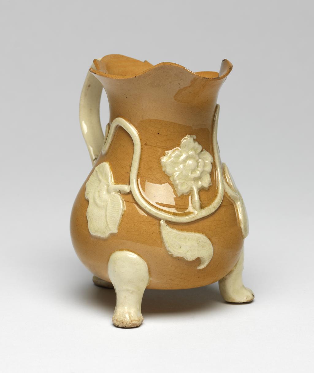 An image of Cream Jug/Milk Jug. Unidentified Staffordshire Pottery. Of silver shape, with pear-shaped body, shaped rim and lip, ear-shaped handle with a kick at the lower end, and three low feet. The front is decorated with an applied wavy stem bearing three moulded leaves and a flower. Ochre-coloured earthenware, the handle, feet and moulded decoration applied in cream clay and lead-glazed. Height 9.2 cm, width 8.5 cm, 1745-1755. Rococo. Sir Ivor and Lady Batchelor Bequest.