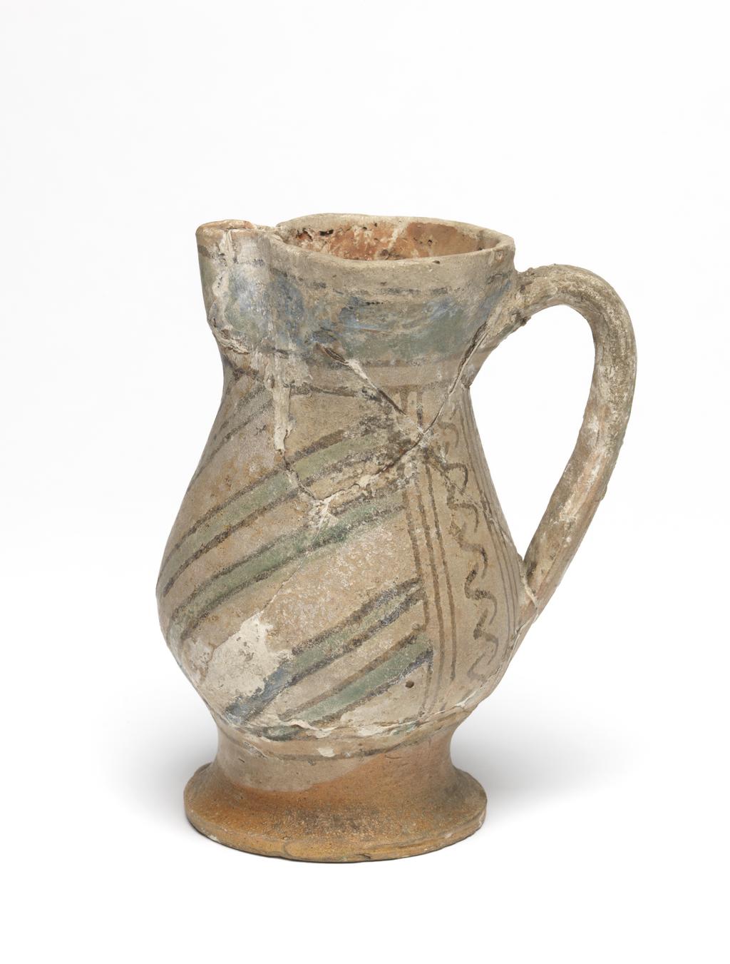 An image of Maiolica. Jug. Unknown maker, probably Italy, Tuscany, Siena. Shape 18. Pear-shaped body standing on a low, slightly spreading foot; the cylindrical neck expands towards the rim which is pinched at the front to form a lip; loop handle of oval section. On each side of the handle, there is a vertical row of Ss between two sets of three black vertical lines. The rest of the body is diagonally striped with green lines edged with black. Above and below are pairs of horizontal black bands, and, on the neck, a broad wavy green band with a black band above. The handle has two groups of horizontal black stripes with three dabs of green between them and a larger dab at its base. Buff earthenware. The interior and foot are lead-glazed yellowish-brown; the rest is tin-glazed pale buff. Painted in black and copper-green. Height, whole, 14.0 cm, diameter, base, 7.0 cm, width, at widest point, 9.4 cm, width, handle to spout, 10.3 cm, circa 1300-1400. Late Medieval.