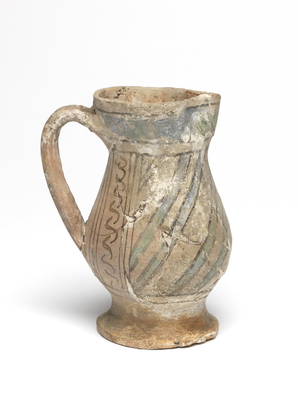 An image of Maiolica. Jug. Unknown maker, probably Italy, Tuscany, Siena. Shape 18. Pear-shaped body standing on a low, slightly spreading foot; the cylindrical neck expands towards the rim which is pinched at the front to form a lip; loop handle of oval section. On each side of the handle, there is a vertical row of Ss between two sets of three black vertical lines. The rest of the body is diagonally striped with green lines edged with black. Above and below are pairs of horizontal black bands, and, on the neck, a broad wavy green band with a black band above. The handle has two groups of horizontal black stripes with three dabs of green between them and a larger dab at its base. Buff earthenware. The interior and foot are lead-glazed yellowish-brown; the rest is tin-glazed pale buff. Painted in black and copper-green. Height, whole, 14.0 cm, diameter, base, 7.0 cm, width, at widest point, 9.4 cm, width, handle to spout, 10.3 cm, circa 1300-1400. Late Medieval.