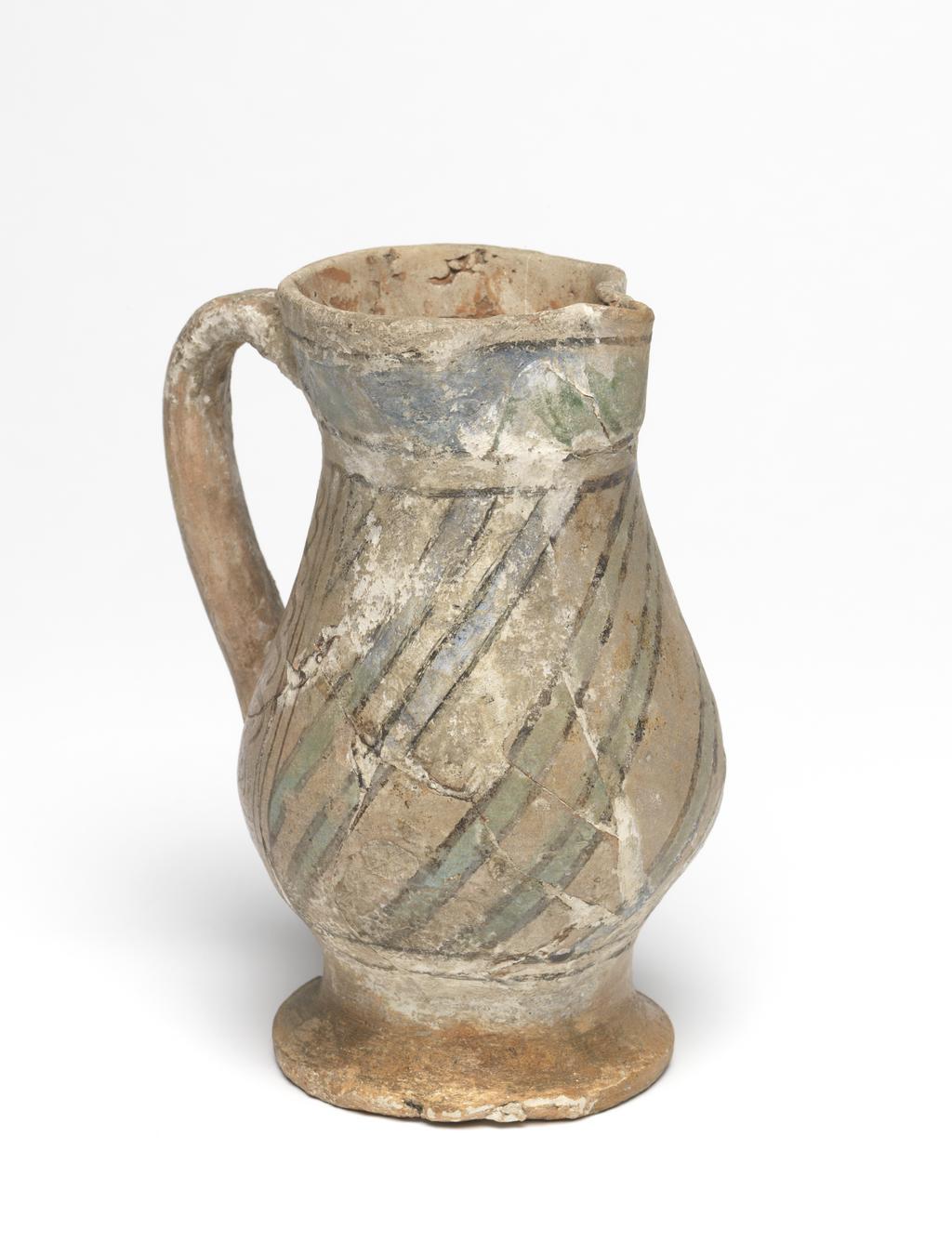 An image of Maiolica. Jug. Unknown maker, probably Italy, Tuscany, Siena. Shape 18. Pear-shaped body standing on a low, slightly spreading foot; the cylindrical neck expands towards the rim which is pinched at the front to form a lip; loop handle of oval section. On each side of the handle, there is a vertical row of Ss between two sets of three black vertical lines. The rest of the body is diagonally striped with green lines edged with black. Above and below are pairs of horizontal black bands, and, on the neck, a broad wavy green band with a black band above. The handle has two groups of horizontal black stripes with three dabs of green between them and a larger dab at its base. Buff earthenware. The interior and foot are lead-glazed yellowish-brown; the rest is tin-glazed pale buff. Painted in black and copper-green. Height, whole, 14.0 cm, diameter, base, 7.0 cm, width, at widest point, 9.4 cm, width, handle to spout, 10.3 cm, circa 1300-1400. Late Medieval.