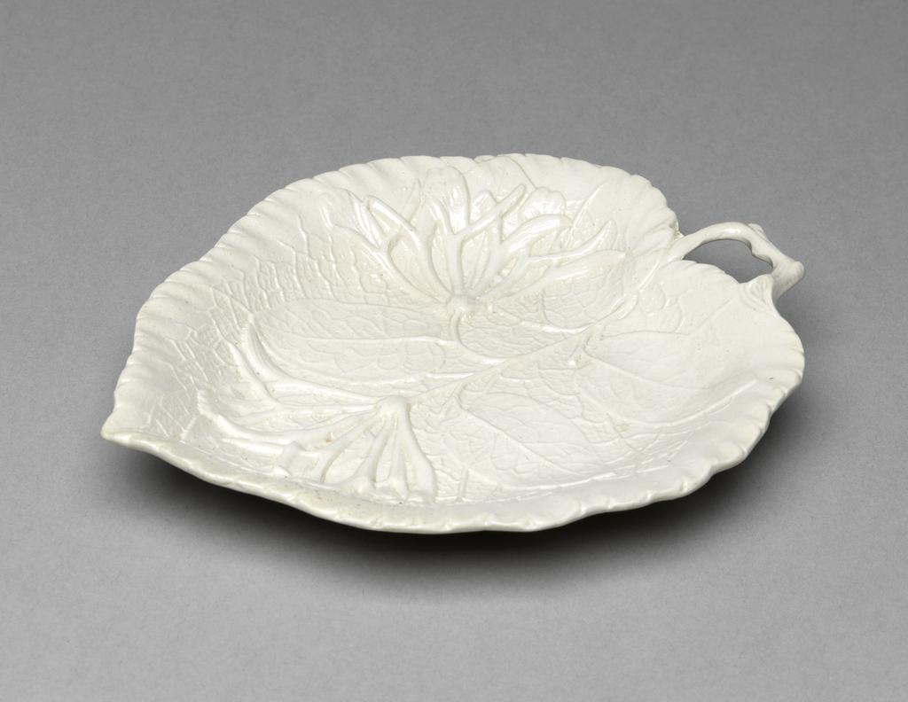 An image of Leaf Dish. Unidentified Staffordshire factory, England. White stoneware, press-moulded in the shape of a veined vine leaf overlaid by a spray of honeysuckle, with a bent twig handle, and three applied fluted flower-shaped feet. 1750-1770. Rococo. Sir Ivor and Lady Batchelor Bequest.