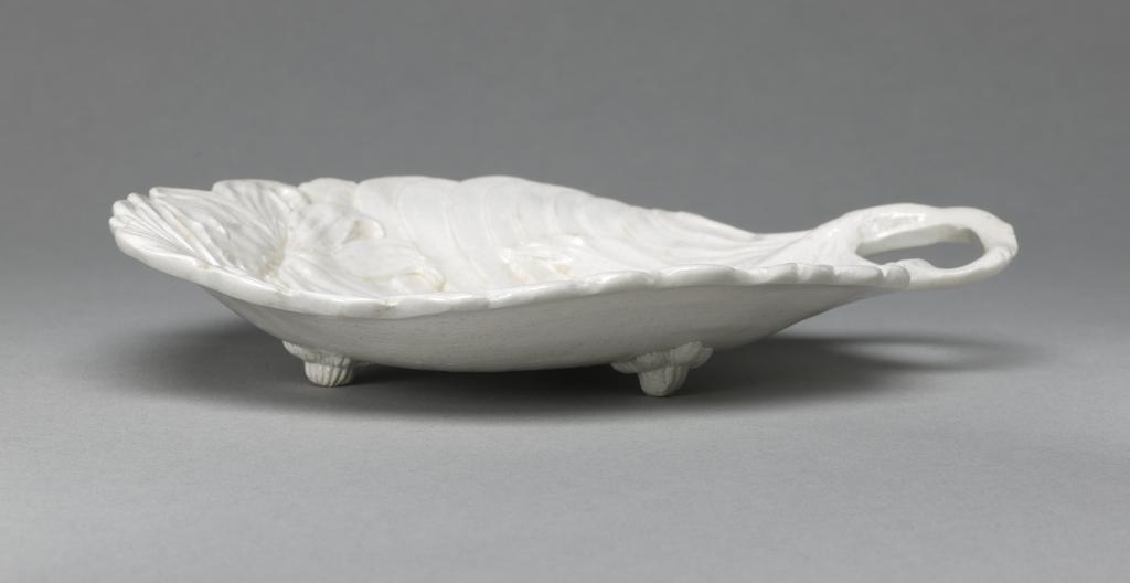 An image of Leaf Dish. Pickle Dish. Unidentified Staffordshire factory, England. In the shape of a concave leaf with a scalloped edge and bent stalk handle, standing on three fluted flower-shaped feet. The interior is decorated with a lily plant with one flower on each side and and three buds at the top, superimposed over the slanting veins of the leaf. Off-white stoneware, press-moulded with integral decoration, and salt-glazed. 1750-1770. Rococo. Sir Ivor and Lady Batchelor Bequest.