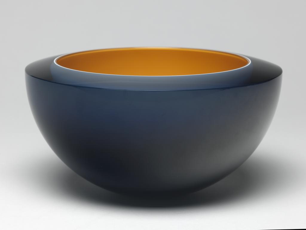 An image of Studio Glass; Contemporary Craft. Bowl with 'floating bowl'. Woodman, Rachael (British, b. 1957). Hemispherical with bevelled top edge, and a small circular base. A dark yellow bowl is cased in a thin layer of white, and a wider layer of translucent dark blue glass, which has been ground away on the base to reveal the white layer. Dark yellow glass, blown and cased in white and blue glass, with bevelled top edge, height, whole, 9.9 cm, diameter, whole, 18.6 cm, dated 1995. Gift of Nicholas and Judith Goodison through the National Art Collections Fund.