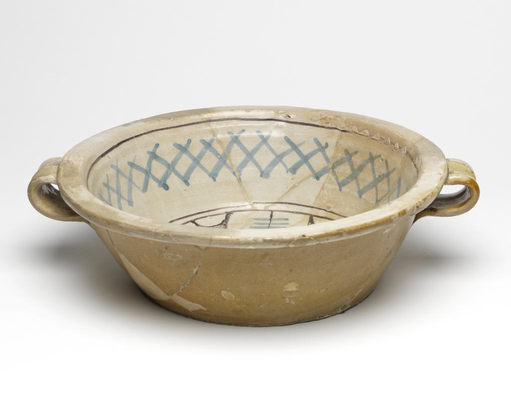An image of Maiolica arcaica blu. Two-handled bowl. Unidentified pottery, probably Umbria, Orvieto or Tuscany. Shape 39. Circular with flat base, sloping sides, narrow flat rim and two wide strap handles with a thumb print at the top. Inside, within two manganese concentric circles, there is a double-armed cross with cross-hatching and three scalloped lines between the arms; on the sides, blue criss-crosses and a manganese band. Pale buff earthenware. The exterior and base are lead-glazed pale brown; the interior is tin-glazed pale grey. Painted in manganese and blue. Height, whole, 9.7 cm, diameter, base, 18.8 cm, width, whole, 34.0 cm, circa 1350-1425. Late Medieval.