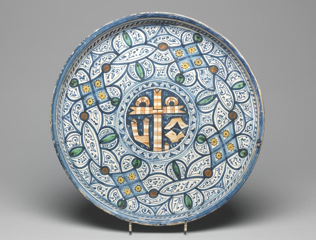 An image of Maiolica. Tray/wall panel. Unknown maker, Tuscany, Montelupo. Circular with shallow vertical sides and flat base which has warped in firing. The central medallion contains a debased form of the Sacred Monogram, outlined in orange and reserved in a blue ground, surrounded by a blue zig-zag border. The rest of the base is decorated with four lozenges with a spot at each point linking them to almond, oval and segmental shapes filled with small strokes and stylised stems and leaves. Round the sides there are short oblique strokes between blue bands and a wide blue band on the rim. The exterior is decorated with three blue horizontal bands. Earthenware, tin-glazed on the front and outside wall and dribbled across the unglazed base. Painted in blue, green, yellow, orange-red, and black. Height, whole, 4.0 cm, diameter, whole, 36.2 cm, circa 1550- 1600. Renaissance. Notes: Blue geometrical decoration comprising rhomboidal and elliptical shapes usually embellished with spots of green, yellow, orange or red has been attributed to Montelupo and Faenza. It seems likely that these bold designs evolved from earlier knot patterns on maiolica, which in turn seem to be related to those on textiles or Islamic metalwork.