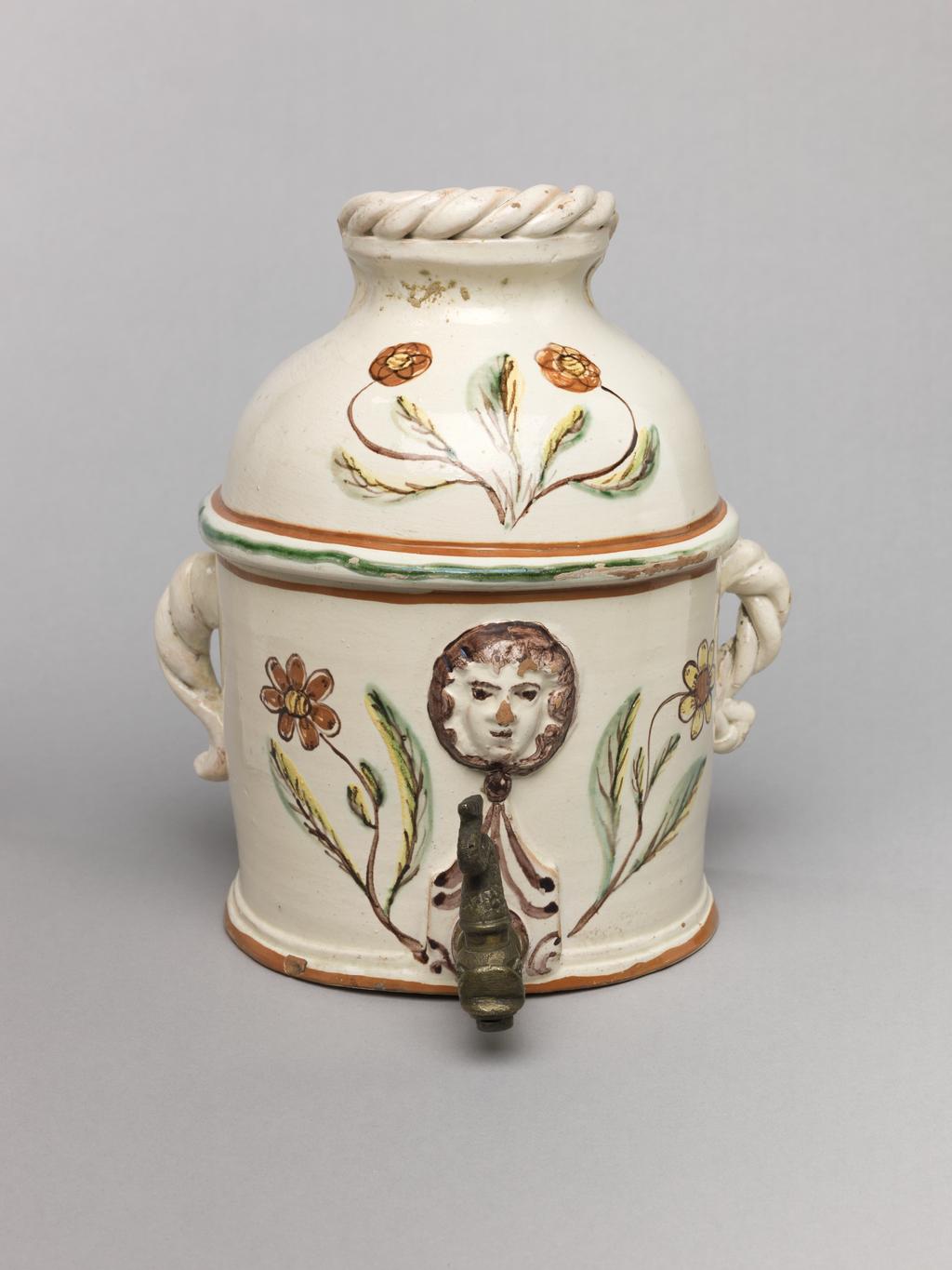 An image of Wall cistern. Lötscher, Andreas (Swiss). Production Place: Switzerland, Canton Graubünden, Sant Antönien. The cistern is semi-circular in plan with straight sides and two twisted handles on opposing sides. The domed top is cut away at the back, and is surmounted by an applied twist. In the middle of the front, a mask in relief is flanked by painted sprays of flowers, and below it, there is a hole in which is inserted a brass tap in the from of a monster's head. Buff earthenware, coated overall except for the back with white slip, and painted in olive-green, red, yellow, and manganese-purple under a clear lead-glaze. Height, whole, 21.5 cm, circa 1823.