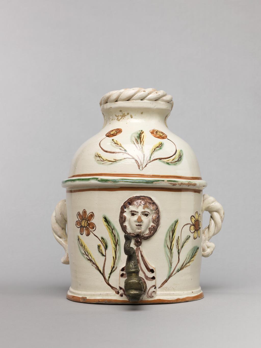 An image of Wall cistern. Lötscher, Andreas (Swiss). Production Place: Switzerland, Canton Graubünden, Sant Antönien. The cistern is semi-circular in plan with straight sides and two twisted handles on opposing sides. The domed top is cut away at the back, and is surmounted by an applied twist. In the middle of the front, a mask in relief is flanked by painted sprays of flowers, and below it, there is a hole in which is inserted a brass tap in the from of a monster's head. Buff earthenware, coated overall except for the back with white slip, and painted in olive-green, red, yellow, and manganese-purple under a clear lead-glaze. Height, whole, 21.5 cm, circa 1823.