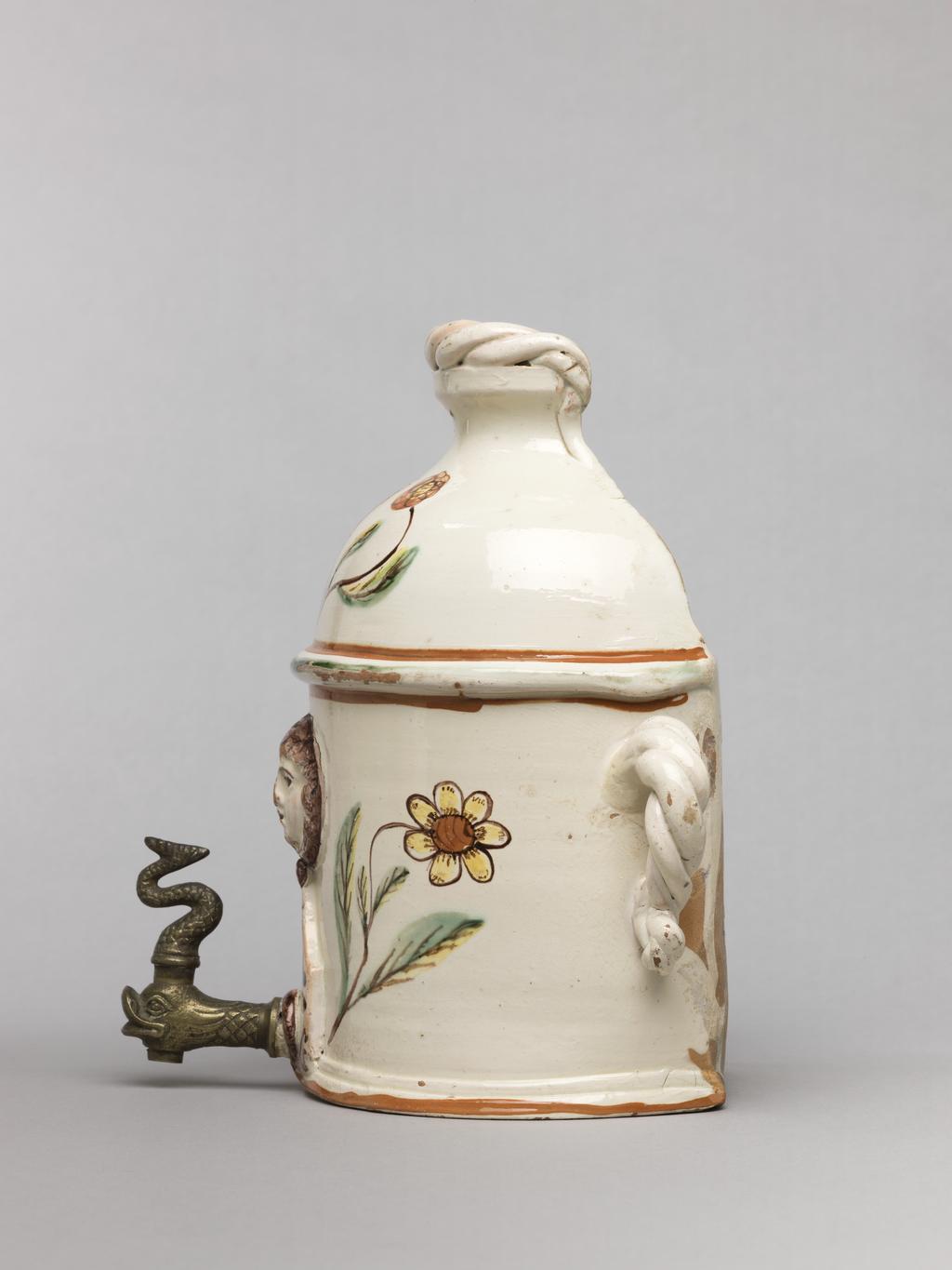 An image of Wall cistern. Lötscher, Andreas (Swiss). Production Place: Switzerland, Canton Graubünden, Sant Antönien. The cistern is semi-circular in plan with straight sides and two twisted handles on opposing sides. The domed top is cut away at the back, and is surmounted by an applied twist. In the middle of the front, a mask in relief is flanked by painted sprays of flowers, and below it, there is a hole in which is inserted a brass tap in the from of a monster's head. Buff earthenware, coated overall except for the back with white slip, and painted in olive-green, red, yellow, and manganese-purple under a clear lead-glaze. Height, whole, 21.5 cm, circa 1823.