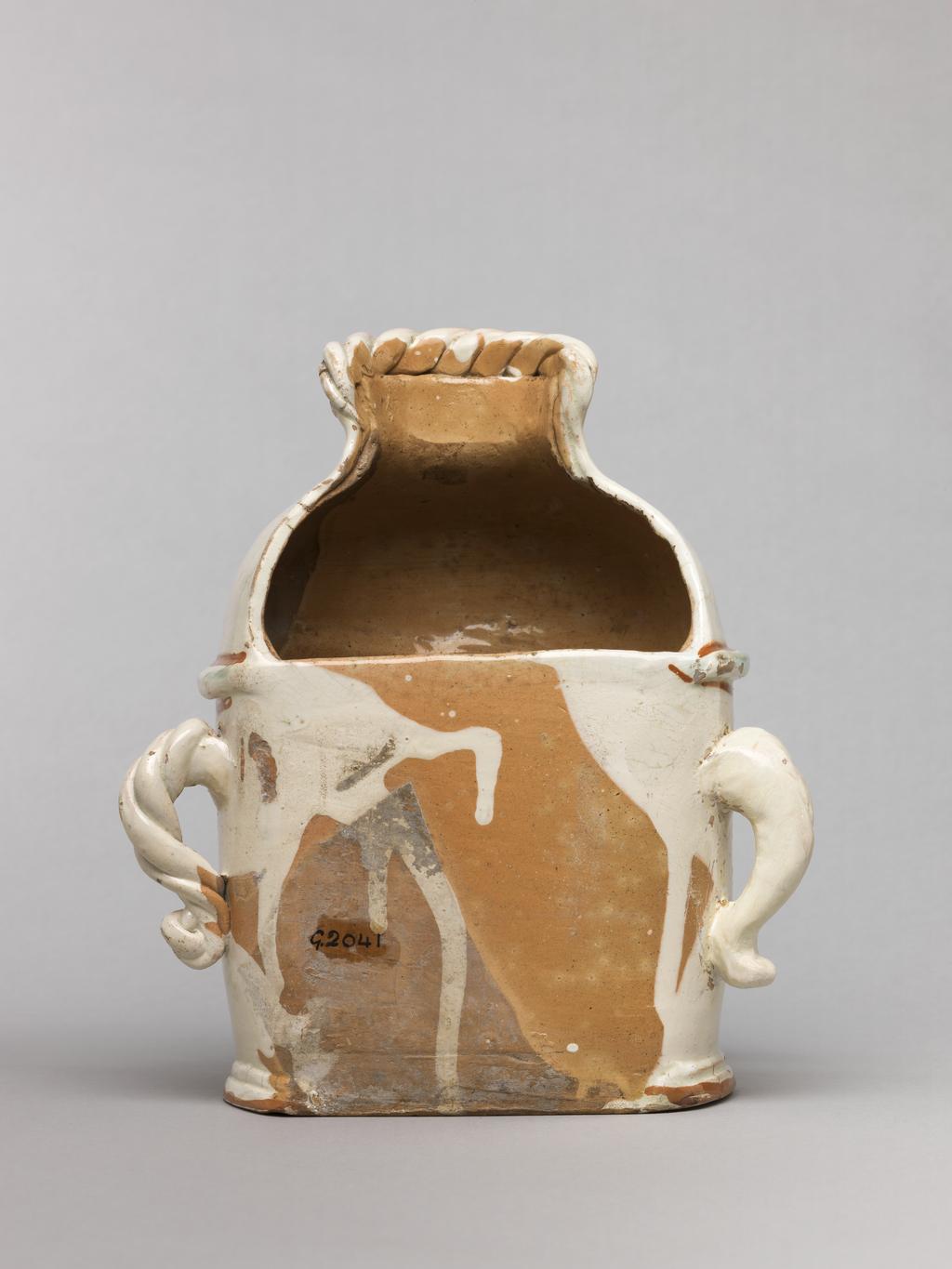 An image of Wall cistern. Lötscher, Andreas (Swiss). Production Place: Switzerland, Canton Graubünden, Sant Antönien. The cistern is semi-circular in plan with straight sides and two twisted handles on opposing sides. The domed top is cut away at the back, and is surmounted by an applied twist. In the middle of the front, a mask in relief is flanked by painted sprays of flowers, and below it, there is a hole in which is inserted a brass tap in the from of a monster's head. Buff earthenware, coated overall except for the back with white slip, and painted in olive-green, red, yellow, and manganese-purple under a clear lead-glaze. Height, whole, 21.5 cm, circa 1823.