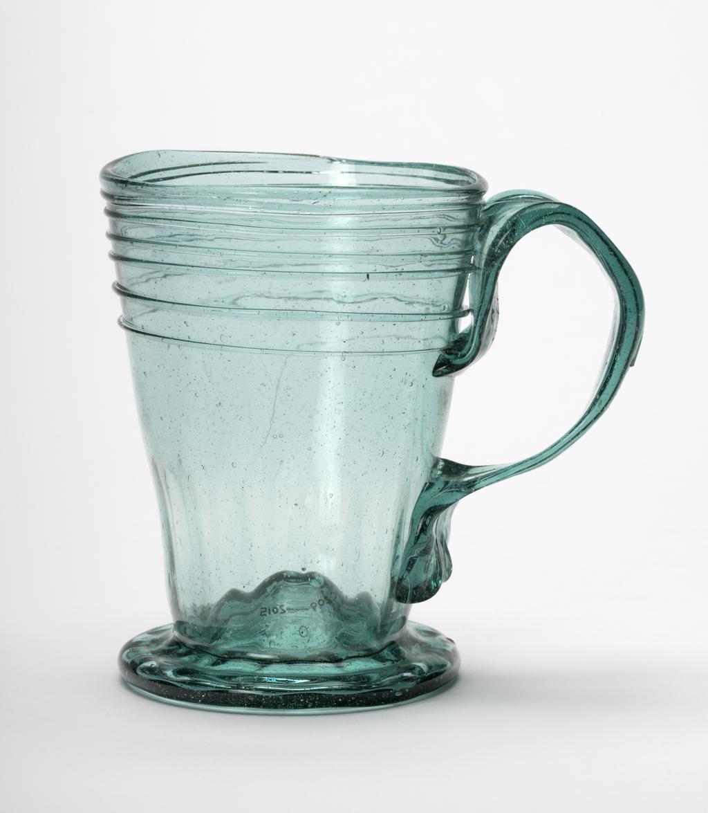 An image of Continental glass. Mug. Unknown glasshouse, Spain, Granada. The foot is dimpled and has a central kick. The sides are lightly fluted on the lower part and have a spirally trailed thread applied to the upper part. The ribbon handle is wider at the top than at the lower end which has a kick pressed back to the handle. Transparent bubbly bluish-green glass, blown. Height, whole, 8.6 cm, width, whole, 10 cm, diameter, rim, 7 cm, circa 1700-1725. Sir Ivor and Lady Batchelor Bequest through The Art Fund.