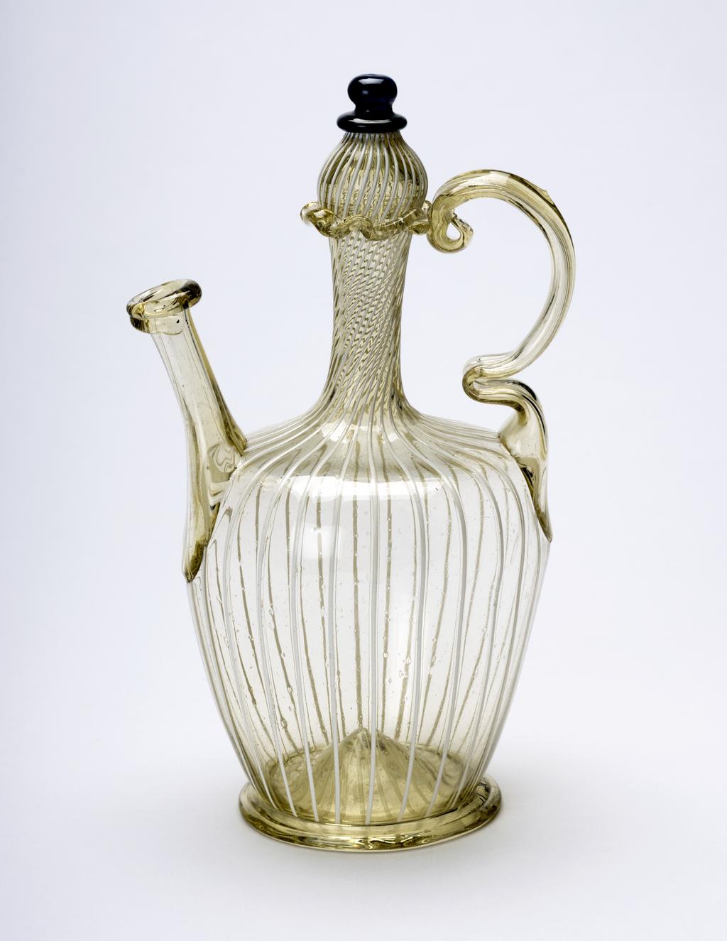 An image of Continental glass. Pharmacy flask. Cruet. Unidentified glasshouse, European. Pale yellowish-green glass with vertical opaque white threads, and dark blue glass finial, height, whole, 16.5 cm, width, whole, 9.6 cm, circa 1600-1799. 18th century Spanish, or 17th century Italian. Production Note: The yellowish-green tone of the glass may indicate that this flask was made in Spain rather than Italy. This form, which is closed except for a small hole in the end of the spout, was used to store liquids which tended to evaporate. A small cover would have been tied over the end of the spout. Sir Ivor and Lady Batchelor Bequest through The Art Fund.
