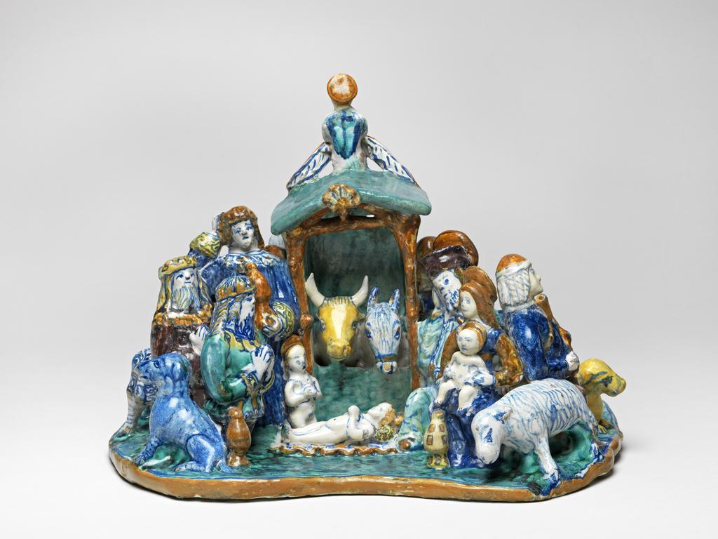An image of Maiolica figure group. The Adoration of the Magi. Giovanni di Nicola di Manzoni of Colle, probably (Tuscany, Colle Val d'Elsa). Formerly attributed doubtfully to Faenza. The Adoration of the Magi modelled in the round on an irregularly shaped base. In the middle is a wattled stall occupied by an ox and an ass. The infant Christ lies on a wavy-edged cloth in front of it, and the infant St John the Baptist kneels at His feet. Further to the left are the three Kings, one standing and two kneeling, accompanied by a dog, who is facing away from Christ. On the right, Joseph kneels behind the Virgin who sits with the infant Christ on her lap. There is a vessel by her feet and, to the right, a sheep. Behind the Virgin, a shepherd holding bagpipes is accompanied by a recumbent dog and two sheep, one resting its forelegs on the back of the other. Behind the three Kings is an attendant with two gaily harnessed horses and a dog. At the back, on the apex of the gable of the stall is a cherub holding a scroll inscribed `O O VOBI. GRADIO.M', and on the wall below is another cherub. On each side there is a kneeling shepherd and a sheep. Buff earthenware, tin-glazed on all the upper surfaces and on the edges of the base. Painted in dark blue, turquoise-green, yellow, orange, and manganese-purple. Height, whole, 25.5 cm, width, whole, 34.5 cm, circa 1509-1515. Renaissance.
