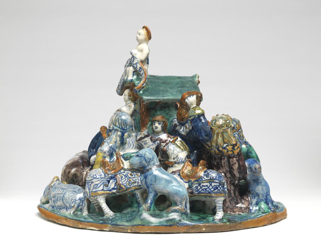 An image of Maiolica figure group. The Adoration of the Magi. Giovanni di Nicola di Manzoni of Colle, probably (Tuscany, Colle Val d'Elsa). Formerly attributed doubtfully to Faenza. The Adoration of the Magi modelled in the round on an irregularly shaped base. In the middle is a wattled stall occupied by an ox and an ass. The infant Christ lies on a wavy-edged cloth in front of it, and the infant St John the Baptist kneels at His feet. Further to the left are the three Kings, one standing and two kneeling, accompanied by a dog, who is facing away from Christ. On the right, Joseph kneels behind the Virgin who sits with the infant Christ on her lap. There is a vessel by her feet and, to the right, a sheep. Behind the Virgin, a shepherd holding bagpipes is accompanied by a recumbent dog and two sheep, one resting its forelegs on the back of the other. Behind the three Kings is an attendant with two gaily harnessed horses and a dog. At the back, on the apex of the gable of the stall is a cherub holding a scroll inscribed `O O VOBI. GRADIO.M', and on the wall below is another cherub. On each side there is a kneeling shepherd and a sheep. Buff earthenware, tin-glazed on all the upper surfaces and on the edges of the base. Painted in dark blue, turquoise-green, yellow, orange, and manganese-purple. Height, whole, 25.5 cm, width, whole, 34.5 cm, circa 1509-1515. Renaissance.