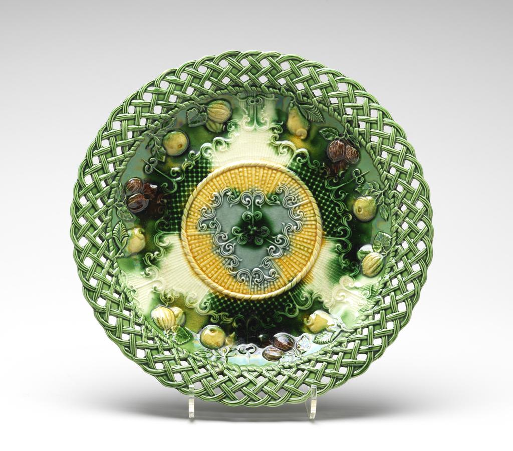 An image of Dessert dish. Unidentified Staffordshire Pottery. Moulded with fruit, nuts, rococo scrolls and diaper, and decorated with coloured glazes. The circular dish has a pierced basket-work border, curved sides, and flat central area. The front is decorated with a central medallion with a yellow rope edging and wickerwork ground enclosing three paired scrolls within a roughly triangular frame of scrolls all in green. On the sides there are three scroll-edged compartments, each enclosing four different fruits and nuts on twigs with leaves suspended from the basket-work border. The space beween the fruits and the central medallion is filled with panels of trellis diaper alternating with panels of striations. Dark cream earthenware, press-moulded with integral relief decoration, pierced, and decorated with green, yellow and manganese-brown coloured glazes. The green glaze does not cover the whole of the area inside the footring, but shows the dark cream body. Height, whole, 3.2 cm, diameter, whole, 28 cm, circa 1755-1765. Rococo. Sir Ivor and Lady Batchelor Bequest through The Art Fund.