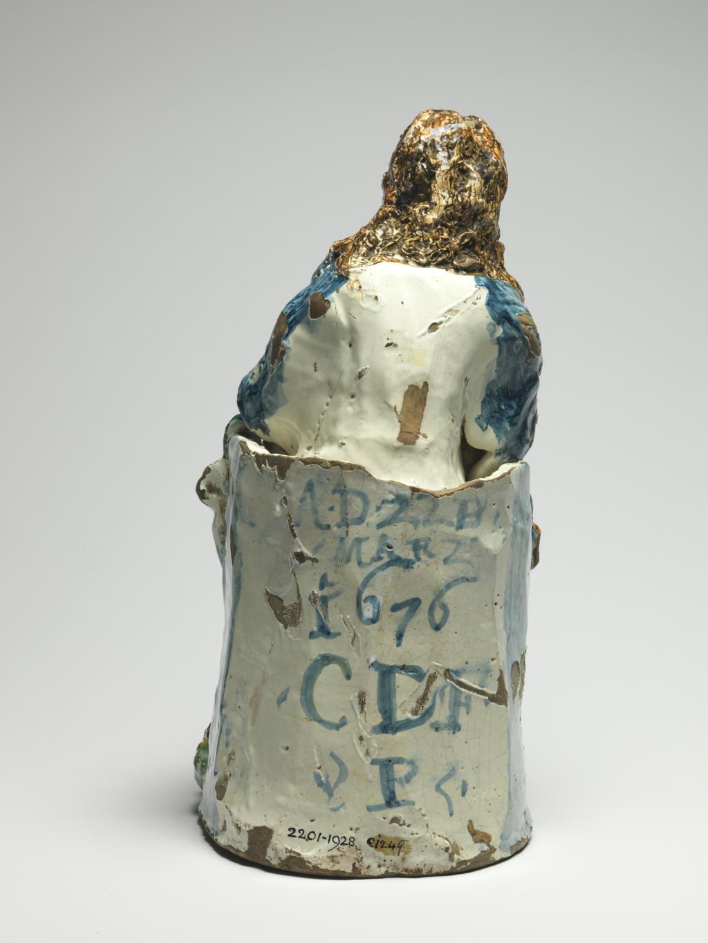 An image of Maiolica. Figure Group. The Virgin and Child enthroned. Unidentified Deruta potter, Umbria. The Virgin sits on a semi-circular throne with a winged cherub's head on each side at the front. She wears a long-sleeved yellow and orange striped gown and a blue cloak scattered with yellow stars. The infant Christ holding an orb sits on her right knee, and she supports a book on her left knee. The green base is decorated along the shaped front edge with three winged cherub's heads in relief. The top is inscribed in manganese `CF/PC/1676 16../S M'. The back of the throne is inscribed in blue `A.D.22D./MARZO/ 1676/.CDF./ P '. Dark buff earthenware, moulded and modelled in the round; tin-glazed on the exterior and on the hollow interior. Painted in dark blue, green, yellow, brownish-orange, and manganese-purple. Height, whole, 38.5 cm, width, base, 18.4 cm, depth, base, 24.0 cm, dated 22 March 1676. Baroque.