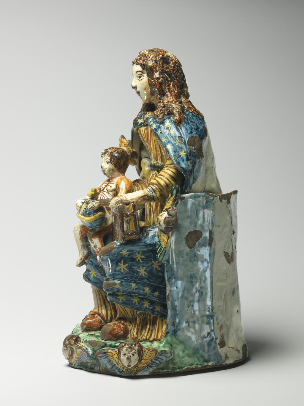 An image of Maiolica. Figure Group. The Virgin and Child enthroned. Unidentified Deruta potter, Umbria. The Virgin sits on a semi-circular throne with a winged cherub's head on each side at the front. She wears a long-sleeved yellow and orange striped gown and a blue cloak scattered with yellow stars. The infant Christ holding an orb sits on her right knee, and she supports a book on her left knee. The green base is decorated along the shaped front edge with three winged cherub's heads in relief. The top is inscribed in manganese `CF/PC/1676 16../S M'. The back of the throne is inscribed in blue `A.D.22D./MARZO/ 1676/.CDF./ P '. Dark buff earthenware, moulded and modelled in the round; tin-glazed on the exterior and on the hollow interior. Painted in dark blue, green, yellow, brownish-orange, and manganese-purple. Height, whole, 38.5 cm, width, base, 18.4 cm, depth, base, 24.0 cm, dated 22 March 1676. Baroque.