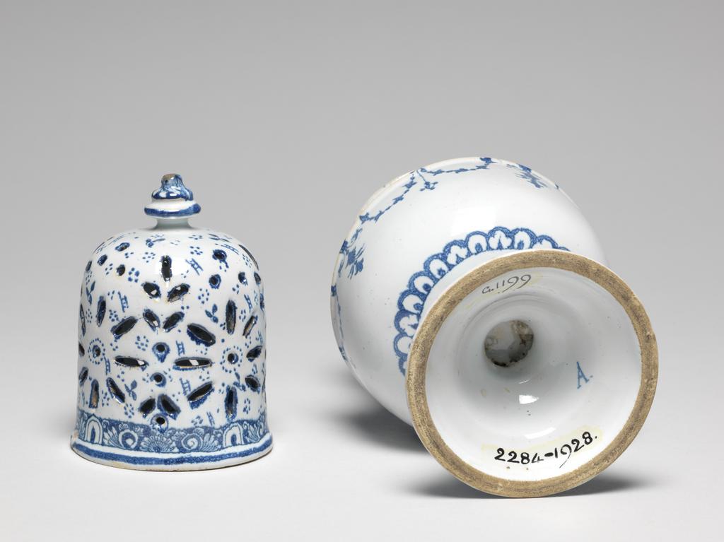 An image of Sugar caster/sugar castor. Unknown factory, France, Rouen. Vase-shaped body supported by a short incurved stem rising from a domed foot of ogee outline. The high, screw-on cover is pierced by petal and leaf motifs, and has a stepped circular finial. The foot and neck have a Bérain style border, that on the neck with pendant swags and tassels below on the shoulder. The cover has a narrow border and the rest of the surface is scattered with groups of dots and small ladder-like motifs beween the openings. The outer edge of the foot and the body around its junction with the stem have a border of stylized leaves. The underside of the foot is marked 'A.' Earthenware, pierced, tin-glazed, and painted in cobalt-blue. Height, whole, 25 cm, diameter, whole, 10.4 cm, diameter, foot, 8.7 cm, circa 1700-1720. Berainesque.