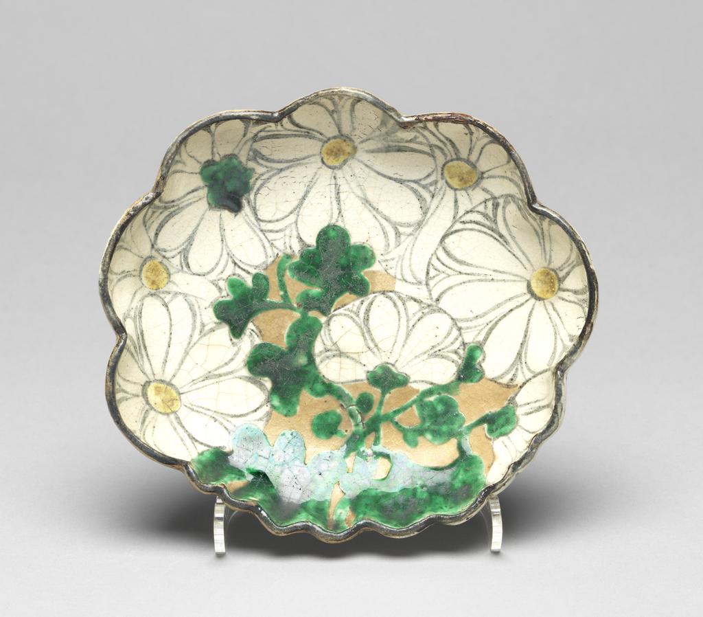 An image of Japanese pottery. Dish. Kenzan, Ogata (1663 - 1743) Japan, Kyoto. Light brown dish of foliate form, moulded with thrown foot. Fawn base glaze, then overglaze decoration with chrysanthemum flowers and foliage in white, yellow and green with black outlines. Some gold background. Stoneware, brush painted, height, whole, 3.30 cm, diameter, dish, 16.60, cm, largest, diameter, foot, 7.90 cm, circa 1600-1743. Edo Period (1615-1868).