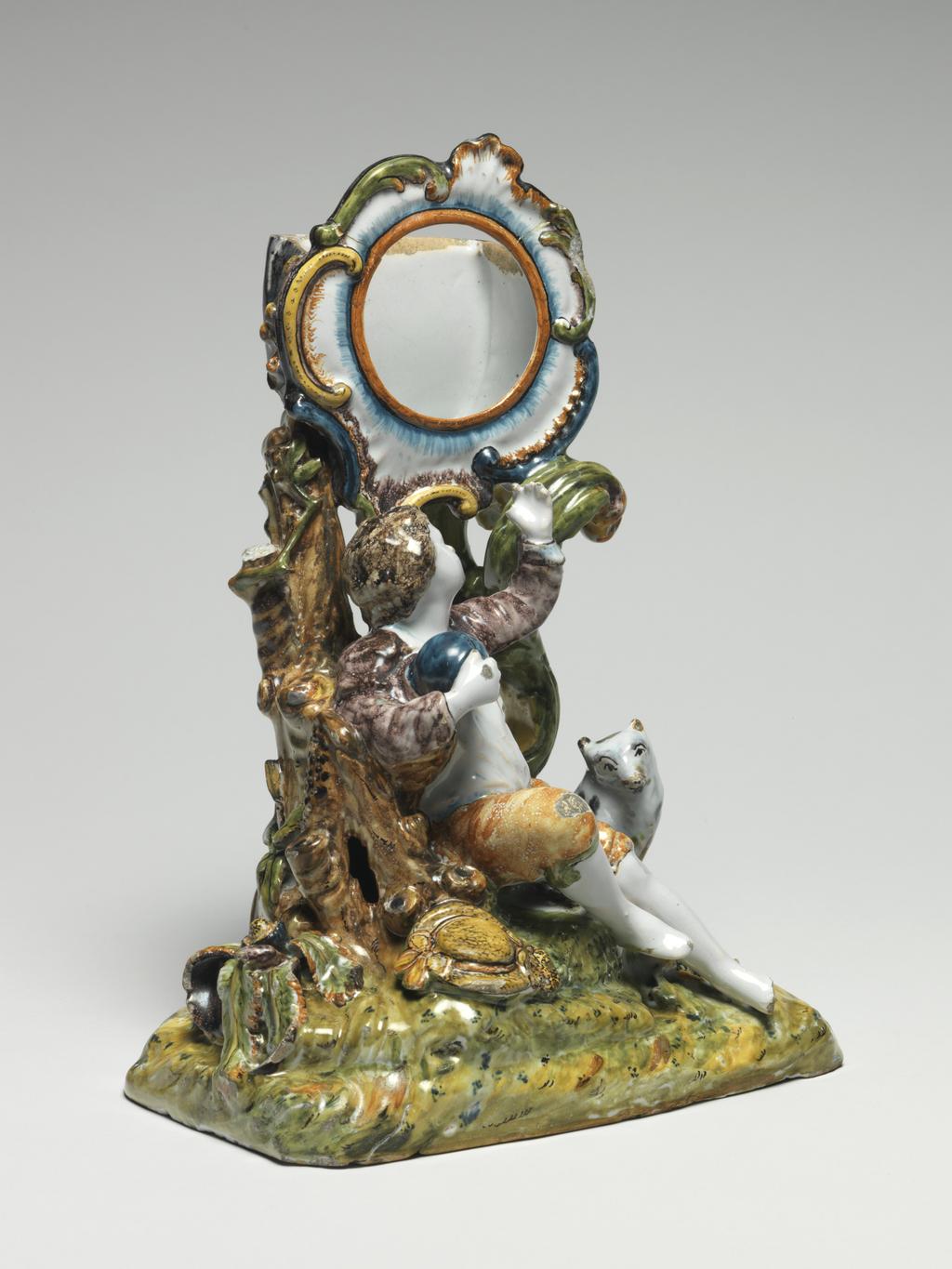 An image of Watch Stand with figures of a Boy and Dog. Unknown, Nièvre, Nevers (formerly in the Nivernais). The rectangular base is open underneath and is decorated on top to resemble a grassy mound. The circular watch holder with a rococo frilled and scrolled edge is supported by a branch of scrollwork and a tree trunk growing upwards from the viewer's left of the base. A barefooted boy holding a blue ball in his right hand and pointing upwards with his left hand sits on the mound with his back to the tree trunk. He wears a white shirt, manganese-purple jacket and pale brown breeches, His yellow broad-brimmed hat trimmed with ribbons lies on the ground to the left. A small white dog with purple and brown patches dog sits to the right of him. Earthenware, moulded in parts, tin-glazed, and painted in blue, olive-green, yellow, manganese-purple, purple, pale yellowish-brown and dark brown high-temperature colours, height, whole, 24.5 cm, width, whole, 17.5 cm, circa 1760. Rococo.
