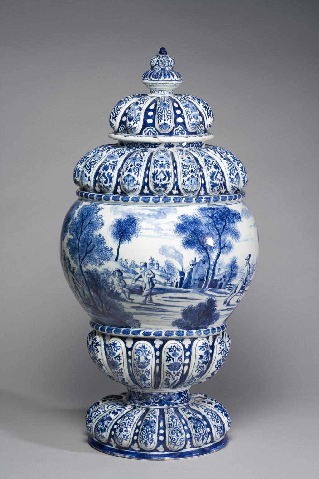 An image of Delftware. Cistern. Unknown Delft factory, Holland. The gadroons are decorated with Chinese-style flowers and symmetrical scrolls of two alternating designs. The convex area is painted with a continuous European landscape with buildings and sponged trees and bushes, amongst which are several men, one of them on a horse, and another smoking a pipe. To one side is a garden with a balustrade and a fountain with dolphins, round which stand four putti. Pale buff earthenware thrown, with applied moulded gadrooning, tin-glazed, and painted in blue, circa 1700. Baroque.