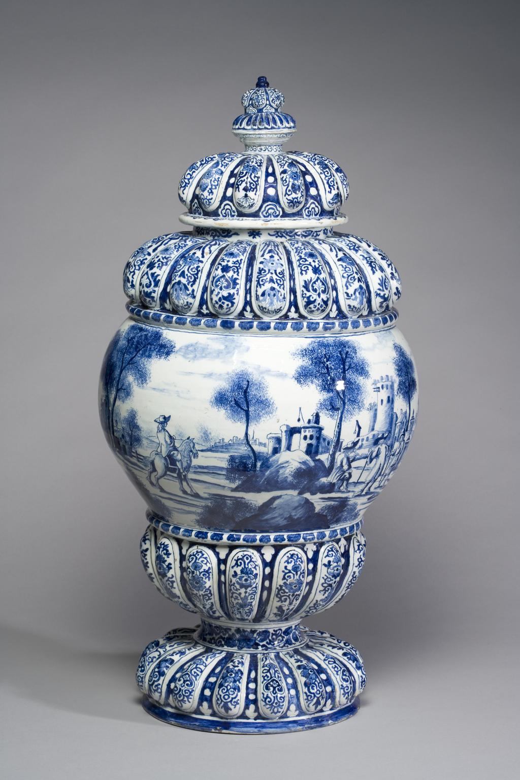 An image of Delftware. Cistern. Unknown Delft factory, Holland. The gadroons are decorated with Chinese-style flowers and symmetrical scrolls of two alternating designs. The convex area is painted with a continuous European landscape with buildings and sponged trees and bushes, amongst which are several men, one of them on a horse, and another smoking a pipe. To one side is a garden with a balustrade and a fountain with dolphins, round which stand four putti. Pale buff earthenware thrown, with applied moulded gadrooning, tin-glazed, and painted in blue, circa 1700. Baroque.