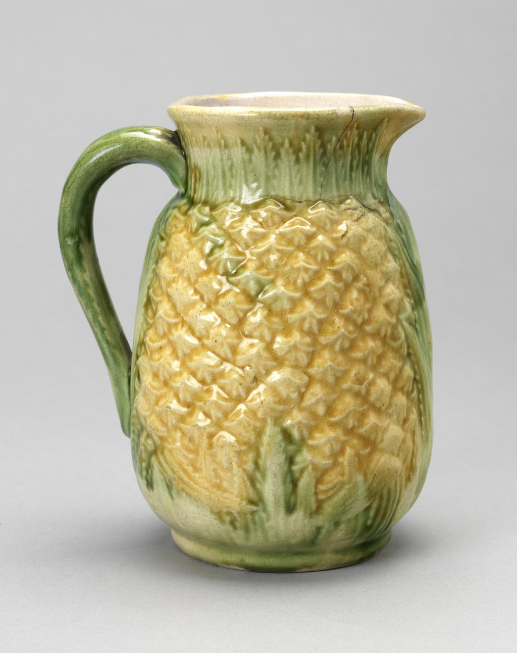 An image of Pineapple jug. Unidentified factory, England. Cream earthenware jug, press moulded in the shape of a pineapple, with a short slightly flaring cylindrical neck, a loop handle and a slim circular foot-rim. The outside is moulded to represent both the fruit and the leaves, the former covered by bright yellow-gold glaze and the latter with a thin green glaze. The interior is tinged pink. Earthenware, press moulded and coated with clear and coloured glazes, height, whole, 14 cm, width, whole, 12 cm, 19th century.