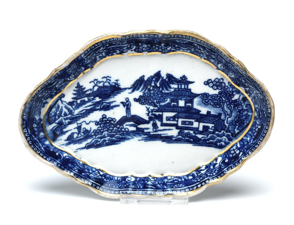 An image of Spoon tray. New Hall Porcelain Factory, England, Staffordshire. Oval with a six-sided shaped edge, shallow, sloping, fluted sides, and flat base. The flat area is decorated with a Chinese landscape, with buildings amid trees on the right, a man near a bridge, and a man standing on a bridge leading to a small island to the left of centre, a boat on water, on the left, a shore with a building amid tree and rocks on the left. On the slides there is border of scrolls, foliage and fruit with dotted beads on the edge. There is a gold band round the lower edge of the sides and round the upper edge. Hybrid hard-paste porcelain printed underglaze in blue, and gilded. Height, whole, 1.4 cm, length, whole, 14.5 cm, width, whole, 9.5 cm, circa 1782-1787. Chinese style.