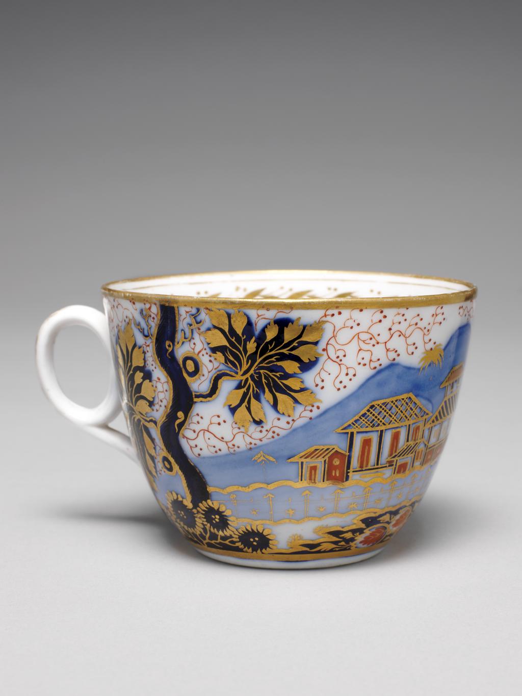 An image of Cup and Saucer/Teacup. New Hall Porcelain Factory, Staffordshire. Bute shape cup with oval ring handle. Circular saucer with curved sides, standing on a footring. The exterior pf the cup is decorated with an Oriental landscape in which are a tree, a group of buildings, a bird perching in a tree branch, and a tiger lying down in front of a rock covered in daisy-like flowers. There is a gold band round the base and rim, inside which is a border of slanting leaves and pairs of berries with a narrow and a wider band above and below. The back of the handle is decorated with five stylized leaves of graduated size. The saucer is decorated with a different version of the design with the tiger lying down on the left below the tree, and with a matching border. Pattern 1214. Bone china, painted underglaze in blue, overglaze in pale apricot and red enamels, and gilded. Circa 1815-1820. Imari style.