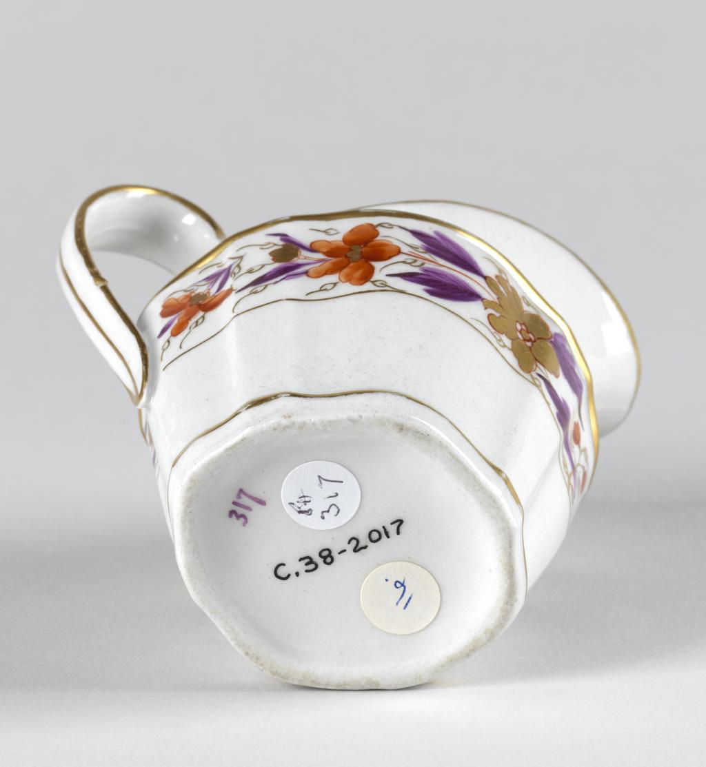An image of Milk jug. New Hall Porcelain Factory, Staffordshire. Pattern no. 317. Of silver-shape with sloping panelled sides, an upward curving lip rising from the shoulder, and high loop handle. Below the shoulder there is a wide border of gold and red flowers with purple leaves with a narrow horizontal band below and a wider band above. On each side of the shoulder there is a gold sprig, and below the lip an arrangement of stylized gold foliage. A gold line runs around the upper edge and down both sides of the handle and another encircles the inside of the upper edge, then joins at the back to run down the centre of the handle. Another gold band encircles the body a little way up from the lower edge. The glaze on the base of the interior is speckled with grey. Hybrid hard-paste porcelain moulded, painted overglaze in red and purple enamels, and gilded. Height, whole, 11 cm, length, whole, 11.6 cm, length, base, 6 cm, circa 1790-1800. Neoclassical.