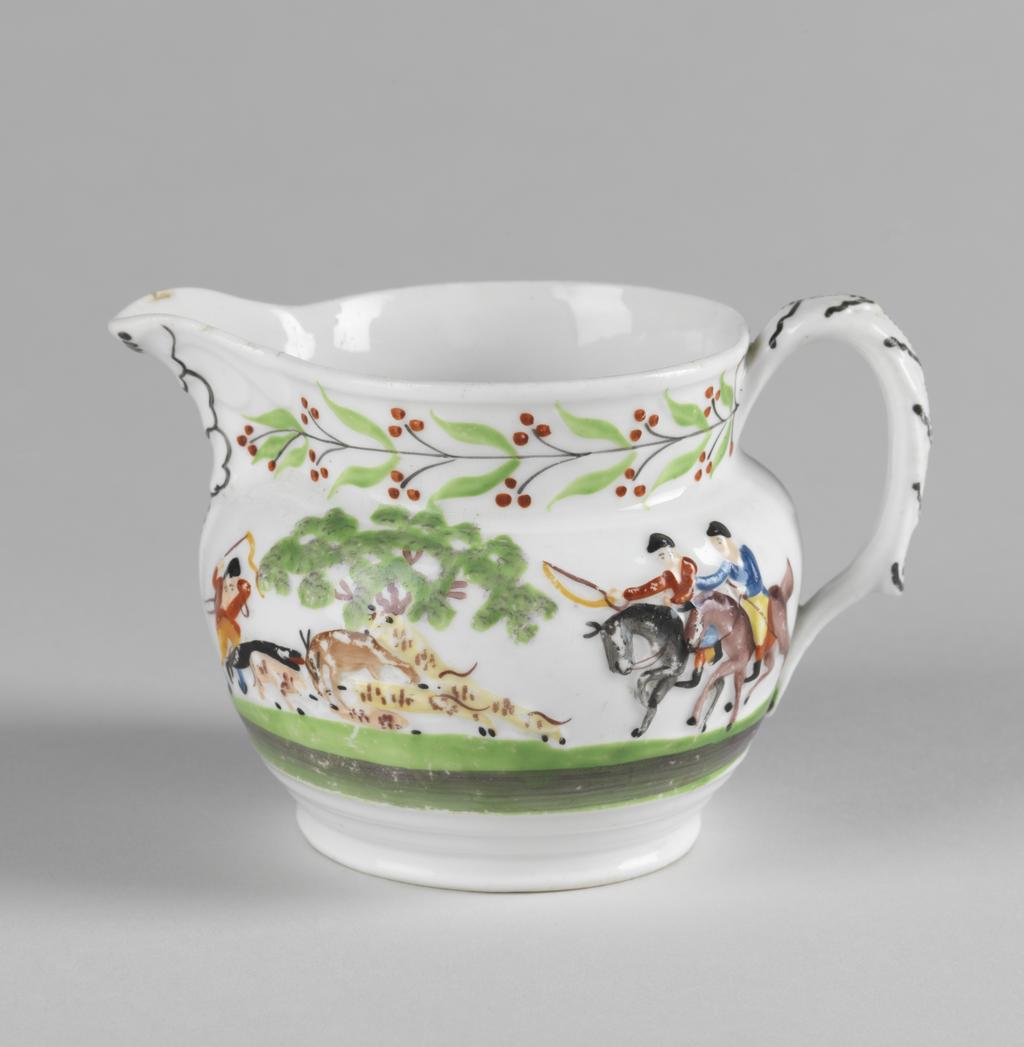 An image of Jug.  New Hall Porcelain Factory, Staffordshire, New Hall. With a hunting scene and a leaf and berry border painted in polychrome enamels. The bulbous jug stands on a low foot, and has a short cylindrical neck with a projecting lip. The strap handle is moulded with leaves in relief, and has a projecting finger rest near the lower end. On the sides, two men on horseback are galloping to the left towards a deer attacked by hounds beside a tree, and another man on foot who is brandishing a whip in one hand and holds the reigns of his horse in the other. On the other side is his horse trotting to the right, and behind it, two trees with a stylized flowering plant between them. Round the neck there is a continuous black stem bearing paired green leaves and paired black stalks with three red berries on each. The leaves on the back of the handle are outlined in black and have black dotted central vein. Bone china, moulded with decoration in relief, painted overglaze in blue, green, yellow, red, brown and black enamels, height, whole, 8.5 cm, length, whole, 12.5 cm, circa 1818-1825.