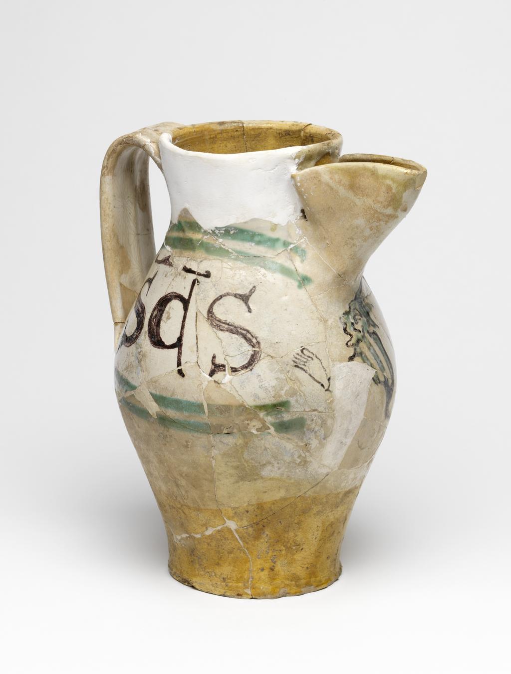 An image of Maiolica arcaica. Jug. Unidentified Orvieto pottery, Italy, Umbria. Shape 7. Ovoid with flat base; cylindrical neck, expanding slightly towards the rim; broad strap handle; spout made from a strip of clay, folded and pressed to the neck. On the front under the spout, a grotesque devil with a striped body, long tail and four horns on its head, faces to the left holding out one of its hands. One side of the jug bears the word `SaS' (Sanctus) in manganese, enclosed by two green lines forming a curved rectangular frame, open on the side nearest the devil. The other side bears the word `IohS' (Johannes), similarly framed. Cream earthenware, the lower part and interior are lead-glazed brownish-yellow, the rest is tin-glazed off-white; the tin-glaze has crazed; painted in manganese and copper-green. Height, whole, 22.7 cm, diameter, base, 8.9 cm, diameter, body, 14.3 cm, width, whole, 17.6 cm, circa 1300-1350. Medieval.