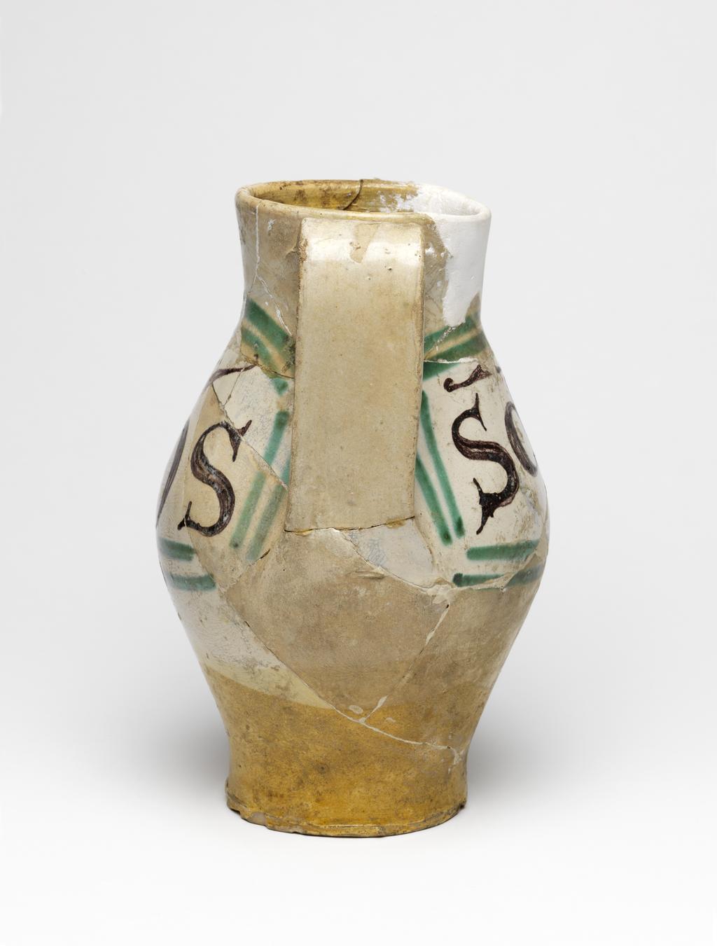An image of Maiolica arcaica. Jug. Unidentified Orvieto pottery, Italy, Umbria. Shape 7. Ovoid with flat base; cylindrical neck, expanding slightly towards the rim; broad strap handle; spout made from a strip of clay, folded and pressed to the neck. On the front under the spout, a grotesque devil with a striped body, long tail and four horns on its head, faces to the left holding out one of its hands. One side of the jug bears the word `SaS' (Sanctus) in manganese, enclosed by two green lines forming a curved rectangular frame, open on the side nearest the devil. The other side bears the word `IohS' (Johannes), similarly framed. Cream earthenware, the lower part and interior are lead-glazed brownish-yellow, the rest is tin-glazed off-white; the tin-glaze has crazed; painted in manganese and copper-green. Height, whole, 22.7 cm, diameter, base, 8.9 cm, diameter, body, 14.3 cm, width, whole, 17.6 cm, circa 1300-1350. Medieval.