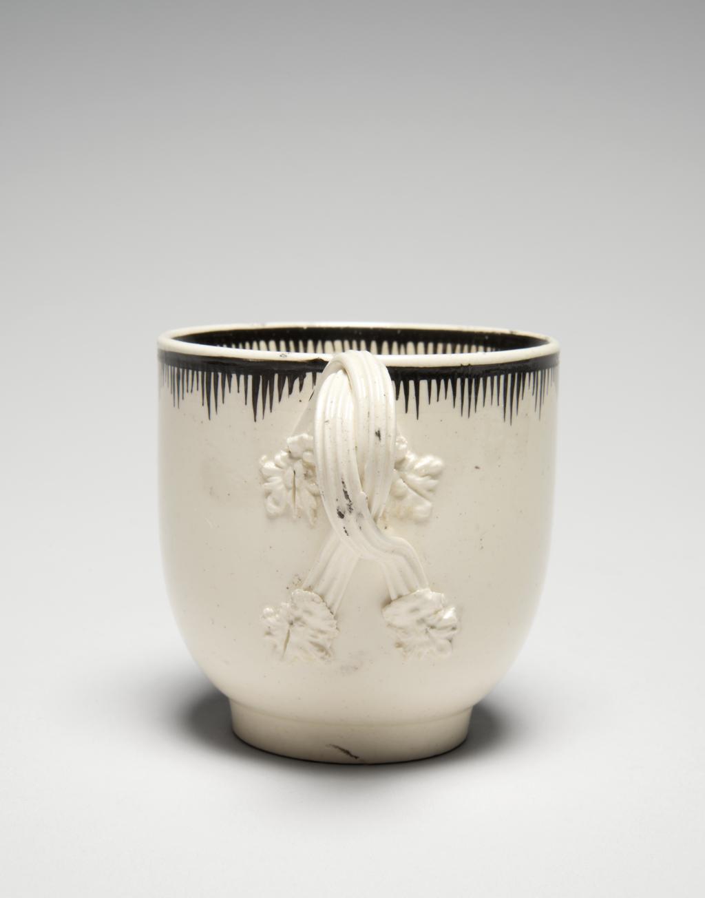 An image of Creamware chocolate cup and saucer, with Minerva and emblems of Wisdom and Industry. Neale & Co., factory, perhaps, Staffordshire. Leeds Pottery, factory, perhaps, Yorkshire. The cup stands on a low footring from which its sides curve outwards and then almost vertically upwards to the rim; reeded double intertwined loop handle with flower and leaf terminals. The saucer has a low footring and deep, concave curving sides. The front of the cup and the centre of the saucer are transfer-printed with a medallion with a medallion enclosing a figure of Minerva, various emblems of Wisdom and Industry and the motto 'LET WISDOM UNITE US', flanked by trophies of game. The interior of the cup is painted with an eight-petalled, stylised flower. Borders on both cup and saucer are composed of a horizontal band from which run close, vertical lines of varying lengths so as to form a rough zig-zag below the band. Cream-coloured earthenware, transfer-printed overglaze and painted in jet black. Pale yellow lead-glaze, inclining towards green where it lies thickly within the footrings. The base and exterior of the cup are lightly crazed with a fine mesh. Height, cup, 6.8 cm, diameter, saucer, 12.6 cm, circa 1780. Rococo.