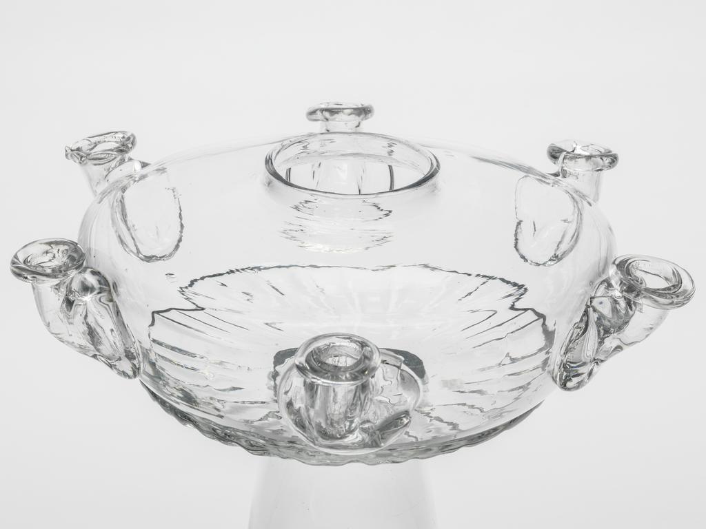 An image of Glass Lamp. Unknown English glassmaker. The depressed globular bowl of the lamp is blown-moulded with gadrooning round the lower part, six applied nozzles for wicks at regular intervals on the sides, and a circular hole in the centre. It is joined to a tall, plain inverted trumpet-shaped foot. Clear lead glass, lamp bowl blown-moulded, nozzles applied decoration, height, whole, 22.0 cm, diameter, foot, 15.7 cm, circa 1700-1725. Baroque. Queen Anne, George I.