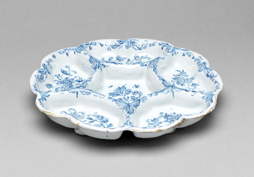 An image of Pickle tray. Sweetmeat dish. Lambeth High Street Pottery, England. Lambeth Griffith, William, proprietor. With six compartments, painted in blue with floral sprays. The glaze on reverse has not adhered to the whole, leaving unglazed areas here and there, The dish is made in six segments with hollows between them on the underside, and stands on five shallow bun feet. The upper side has a central five-sided compartment surrounded by five compartments with three slightly curved internal sides and a two scallops on the outer edge (ten in all). Each compartment is decorated with a different stylized floral spray and the outer edge and raised edges between the compartments have borders of wavy sprays of leaves and stylized flowers. Buff earthenware, press-moulded, with applied feet, tin-glazed pale greyish-white, and painted in blue. Height, whole, 3.4 cm, diameter, whole, 22.3 cm, circa 1750-1760. Sir Ivor and Lady Batchelor Bequest through The Art Fund.