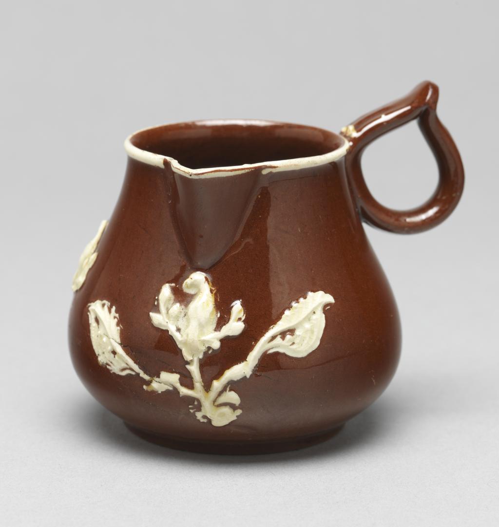 An image of Cream jug/milk jug. Unidentified Staffordshire Pottery, England. The bulbous body rises into a short cylindrical neck with a sparrow beak lip, and at right angles to it, a ring handle pinched together at the top. Below the lip is a sprig of bird perching on an upright branch with a large leaf on either side. Further to the left is a smaller bird perching on a branch with leaves, and to the left of that, a fleur-de-lys. A white slip band runs round the rim. Red earthenware, thrown, with applied spout and handle, decorated with mould-applied white sprigs (reliefs) and white slip under lead-glaze. Height, whole, 7 cm, width, whole, 8.4 cm, circa 1740-1750. Rococo. Sir Ivor and Lady Batchelor Bequest.