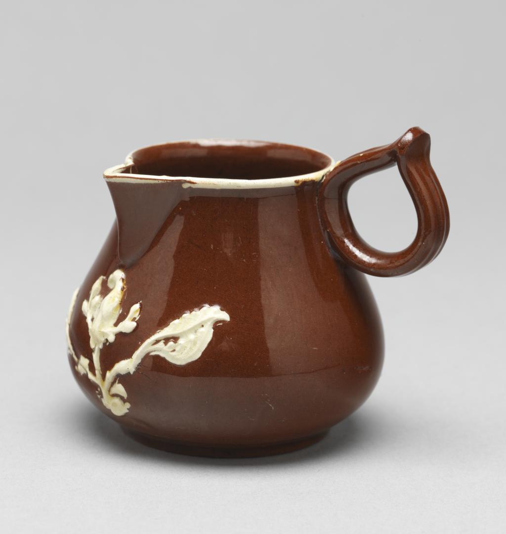 An image of Cream jug/milk jug. Unidentified Staffordshire Pottery, England. The bulbous body rises into a short cylindrical neck with a sparrow beak lip, and at right angles to it, a ring handle pinched together at the top. Below the lip is a sprig of bird perching on an upright branch with a large leaf on either side. Further to the left is a smaller bird perching on a branch with leaves, and to the left of that, a fleur-de-lys. A white slip band runs round the rim. Red earthenware, thrown, with applied spout and handle, decorated with mould-applied white sprigs (reliefs) and white slip under lead-glaze. Height, whole, 7 cm, width, whole, 8.4 cm, circa 1740-1750. Rococo. Sir Ivor and Lady Batchelor Bequest.